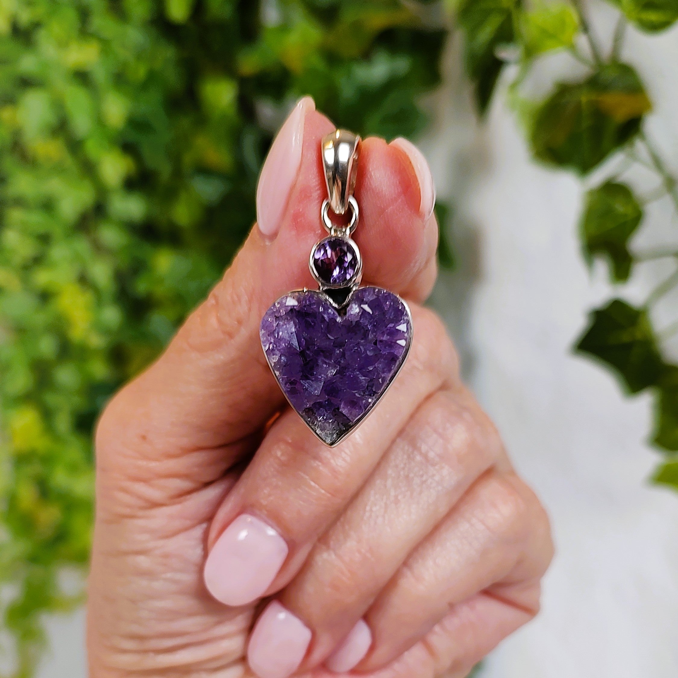 Hand holding a heart-shaped amethyst cluster pendant with a blurred green and white background.