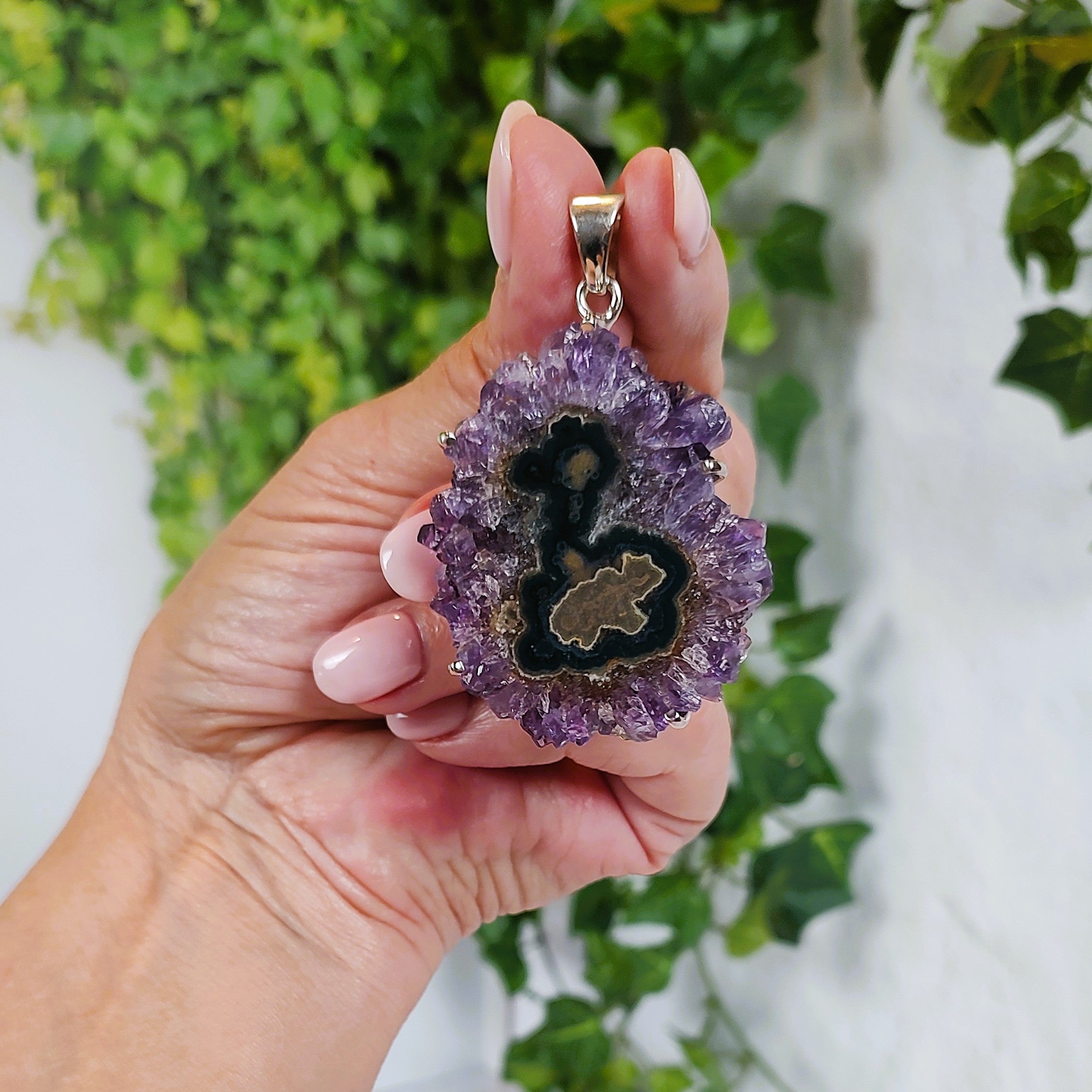 Hand holding an amethyst stalactite slice pendant with green agate in the center, against a green leafy background.