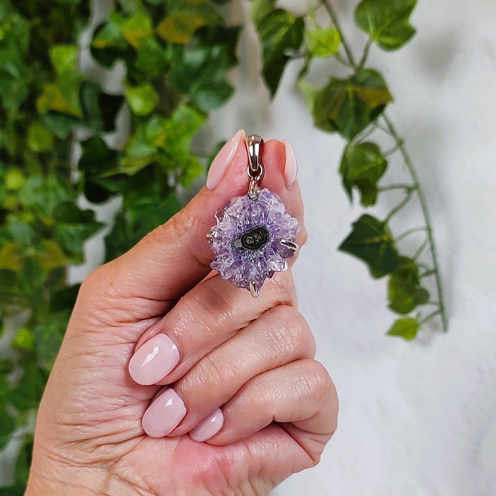 Hand holding an amethyst stalactite pendant with a green agate center, against a background of green leaves.