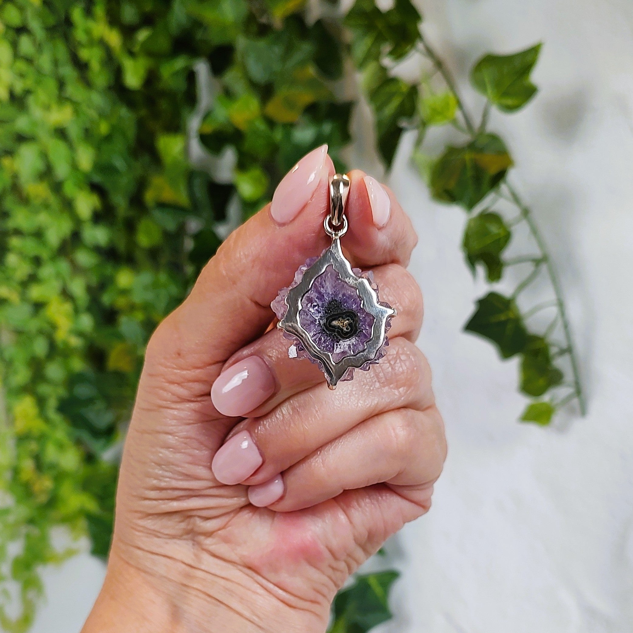 Hand holding an amethyst stalactite pendant with green agate center, showing the back of the pendant, with greenery in the background.