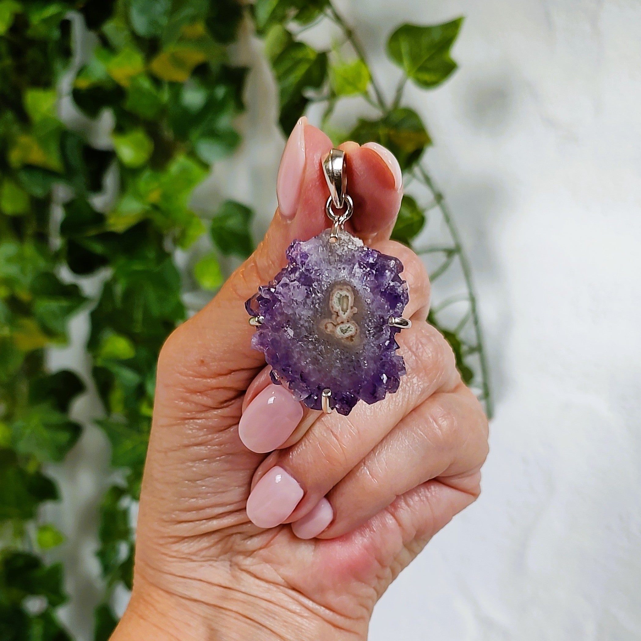 Hand holding an amethyst stalactite slice pendant with a blurred green plant background.