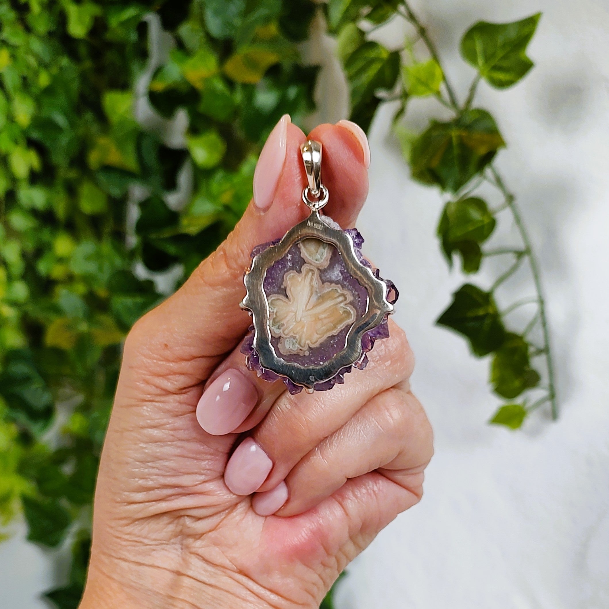 Hand holding an amethyst and agate stalactite pendant, showing the back, against a blurred green leafy background.