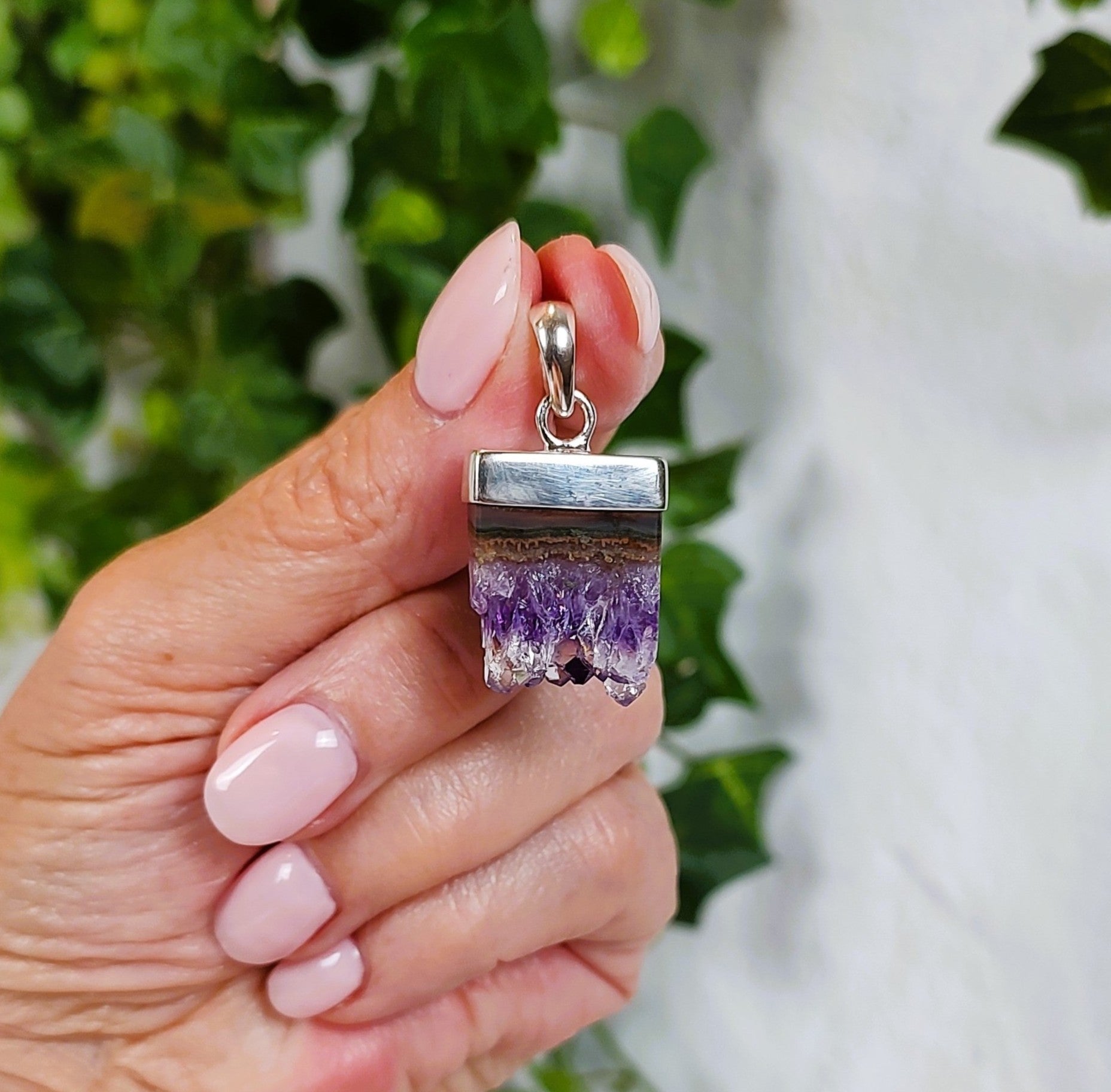 Hand holding a silver pendant with an amethyst and agate stalactite slice, against a blurred green and white background.
