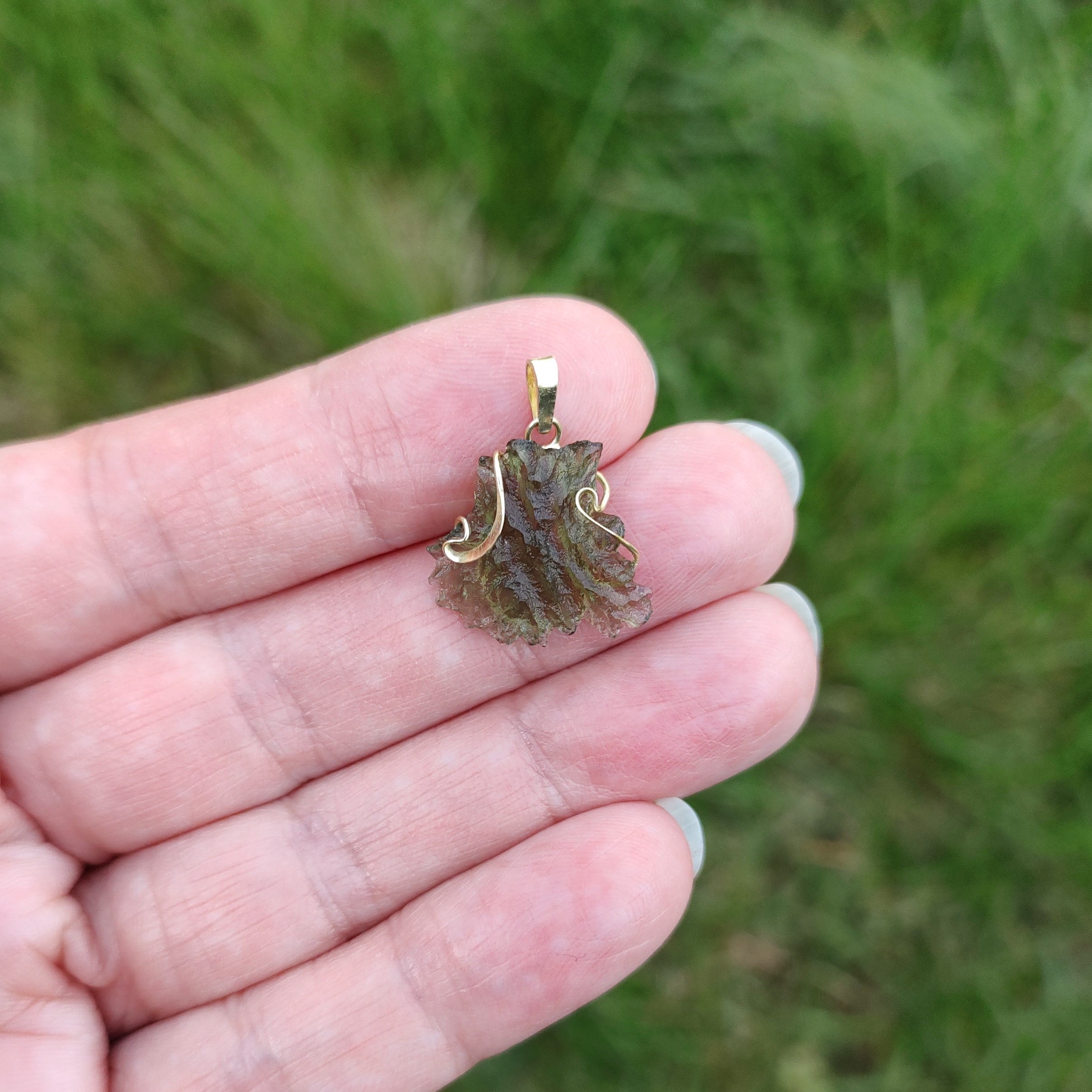 Hand holding a raw moldavite pendant set in gold, against a grassy background.
