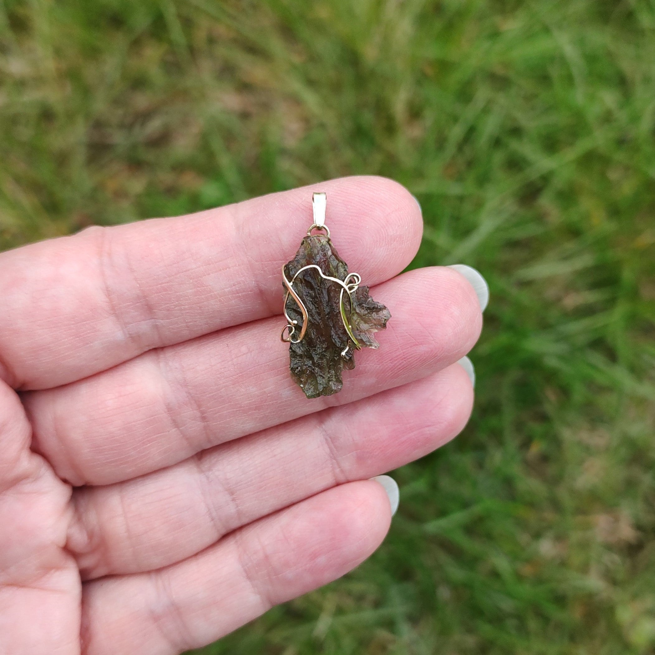 Hand holding a raw moldavite pendant set in gold, against a grassy background.