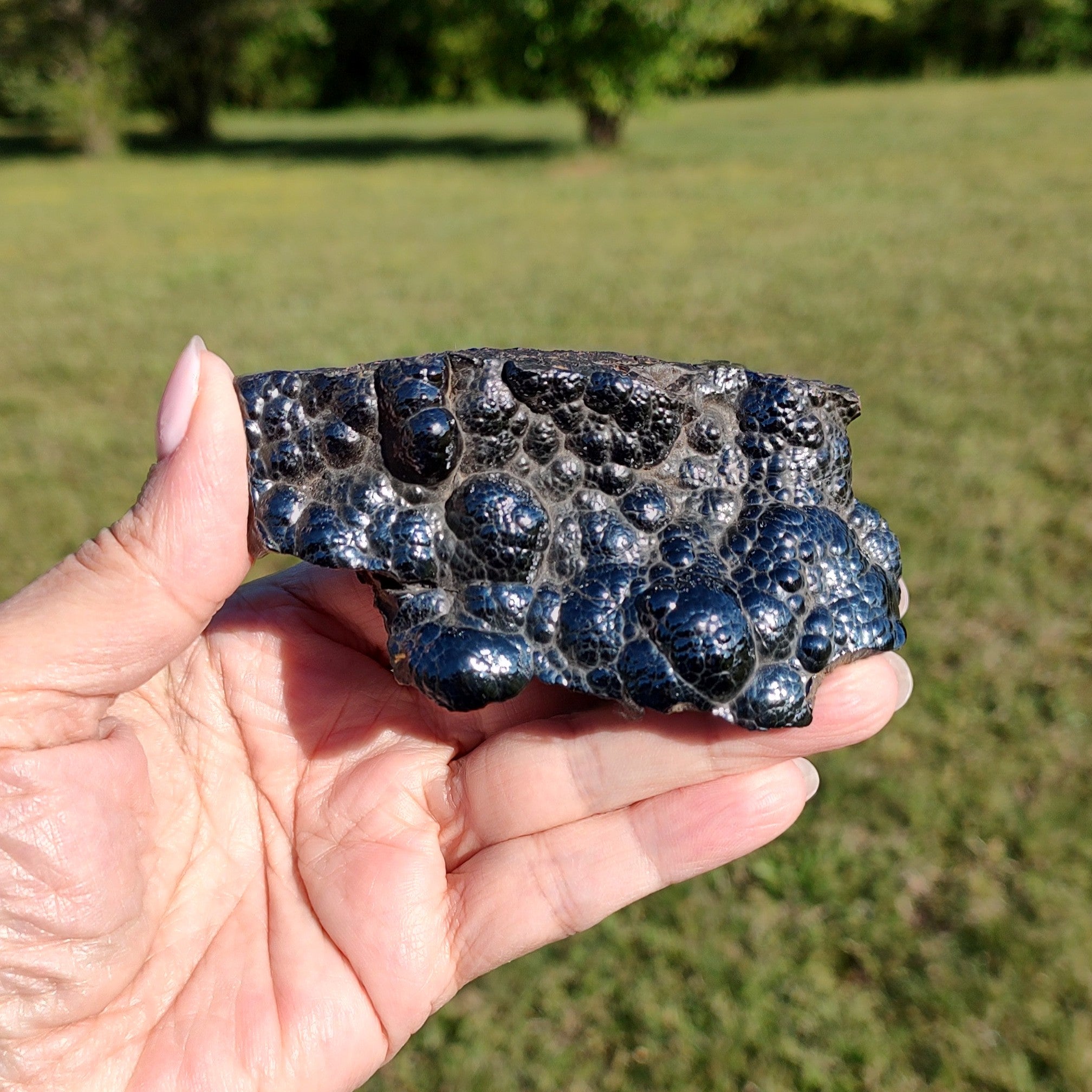 Hand holding a raw hematite specimen against a grassy outdoor background.