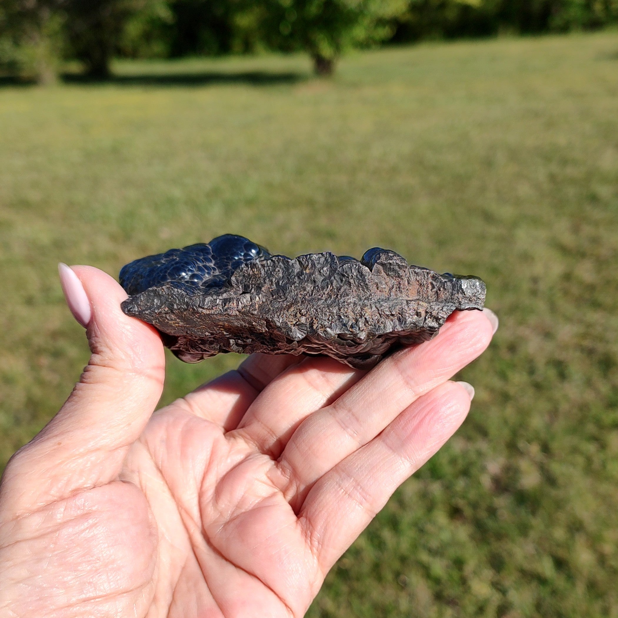 Hand holding a raw hematite specimen, outdoors in front of a grassy field.