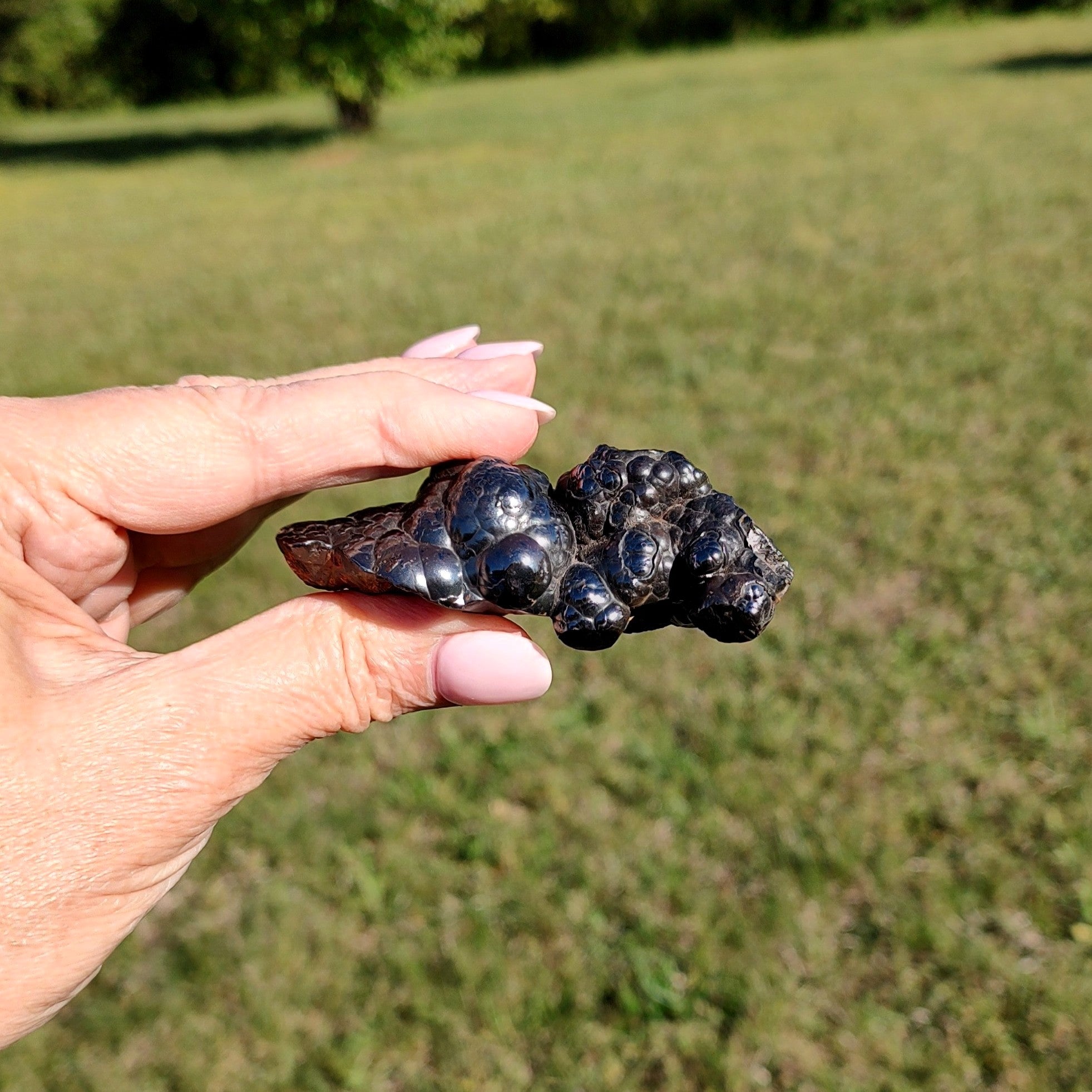 Hand holding a raw specimen of hematite against a blurred green outdoor background.
