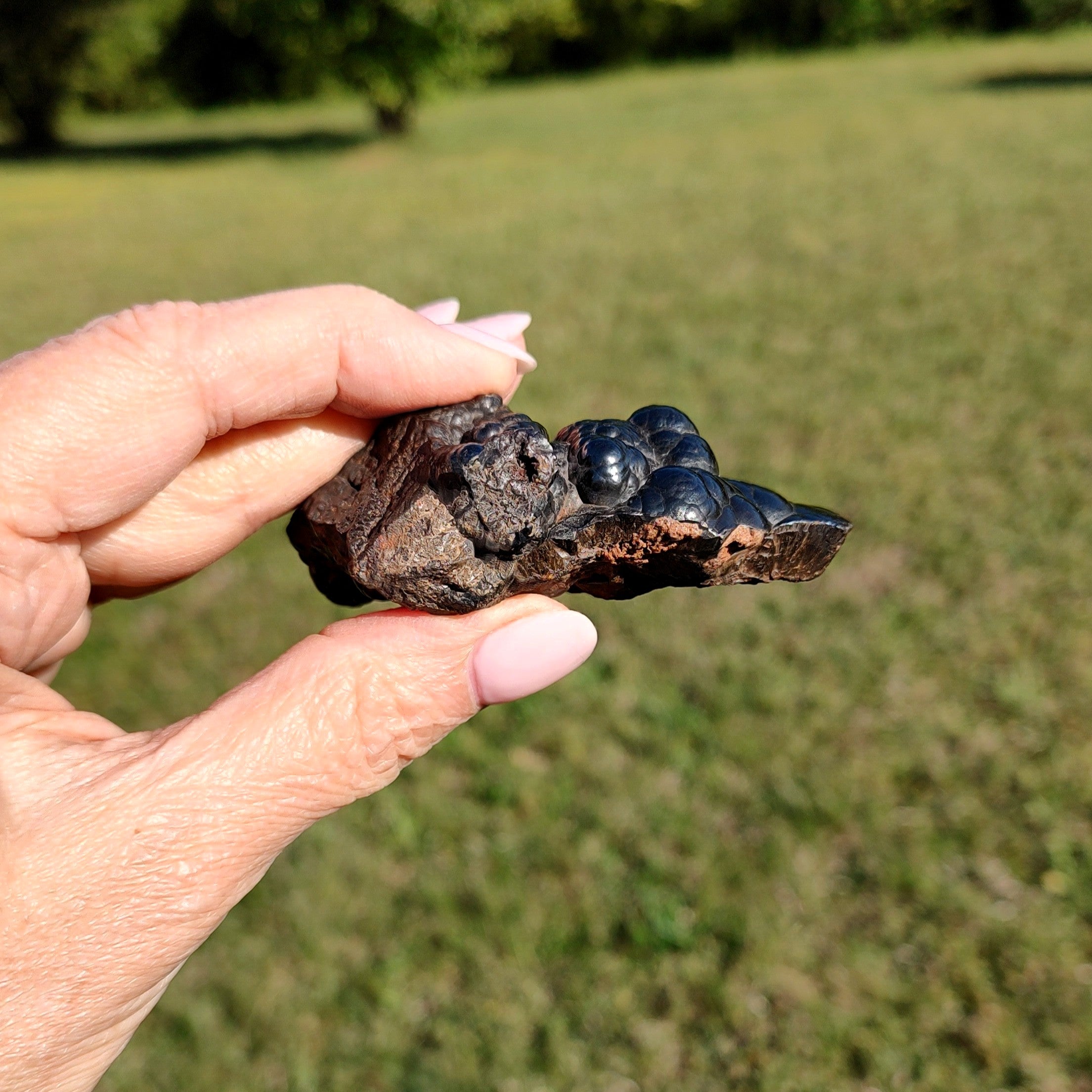 Hand holding a raw hematite specimen in an outdoor setting with green grass.