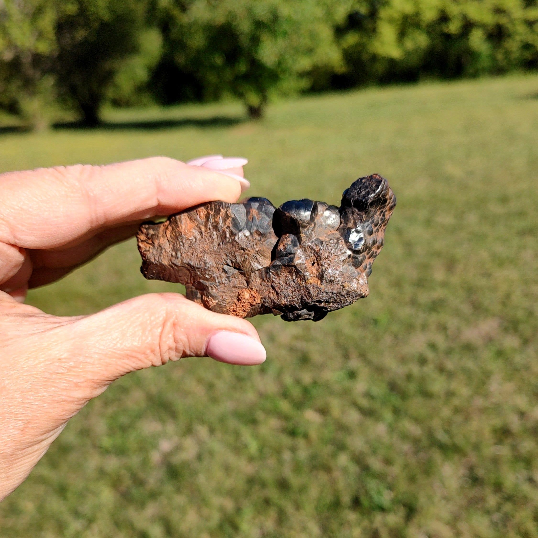 Hand holding a raw hematite specimen with a natural outdoor background.