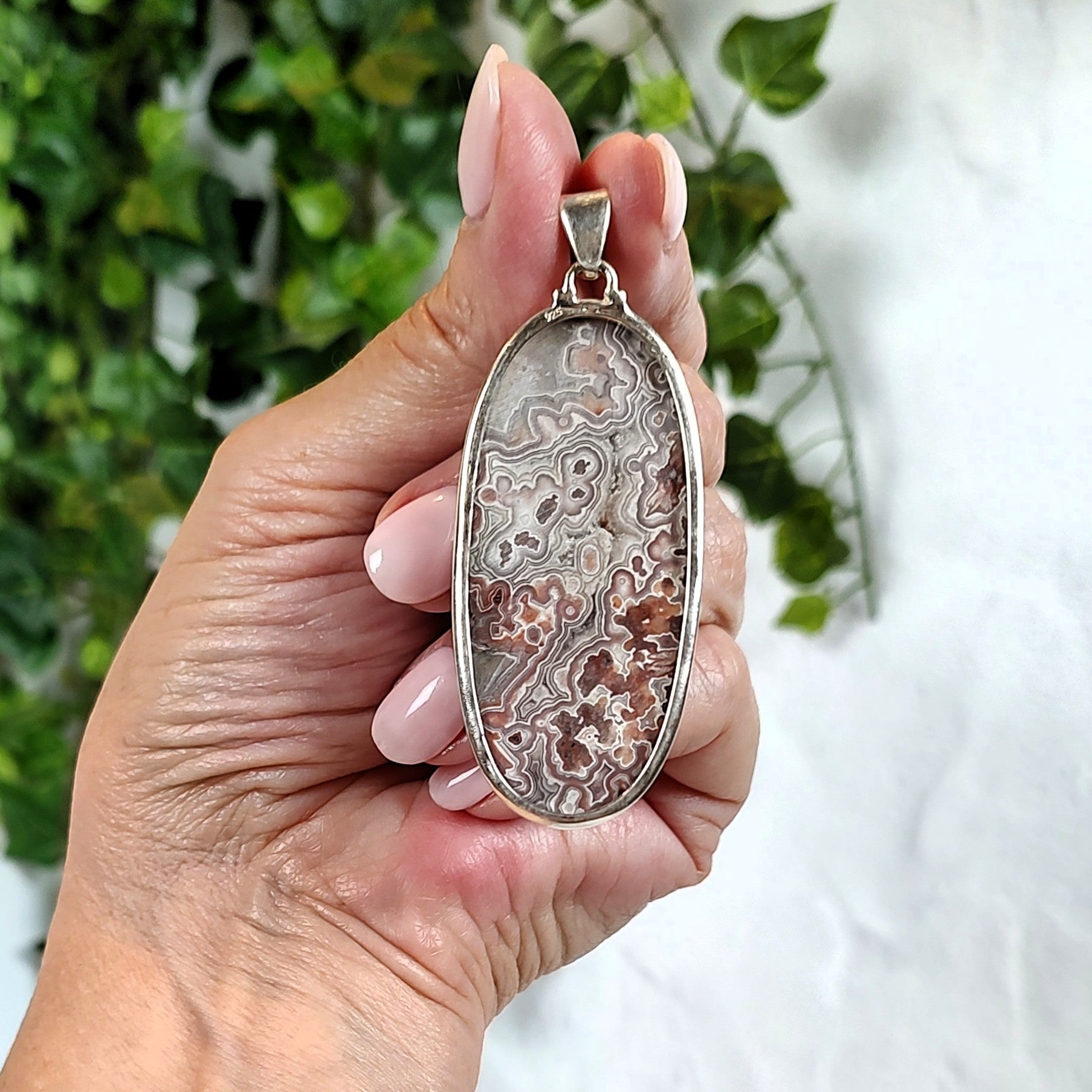 Hand holding a silver pendant set with an agate stone, against a blurred green and white background.