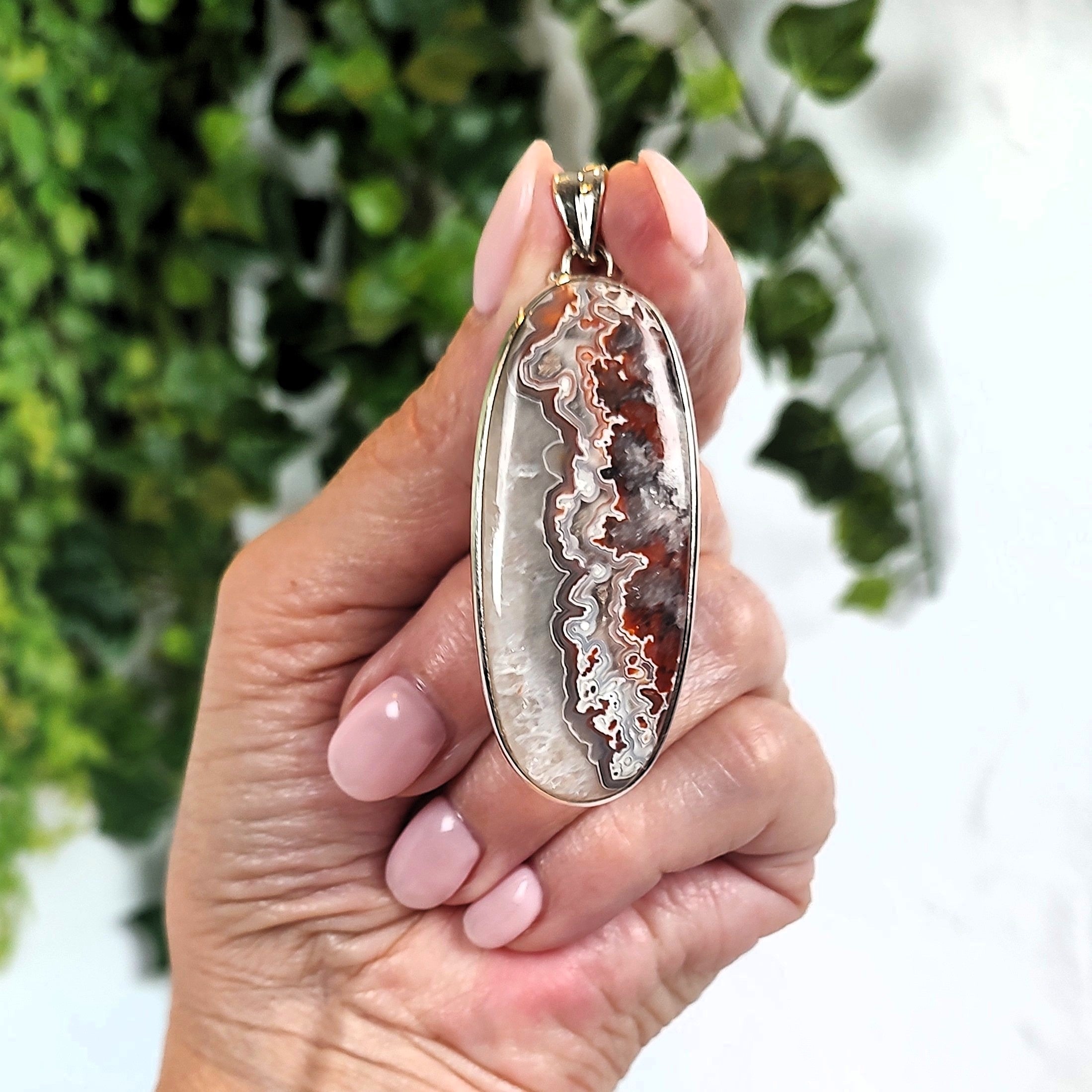 Hand holding a pendant set with an agate stone, against a blurred green and white background.