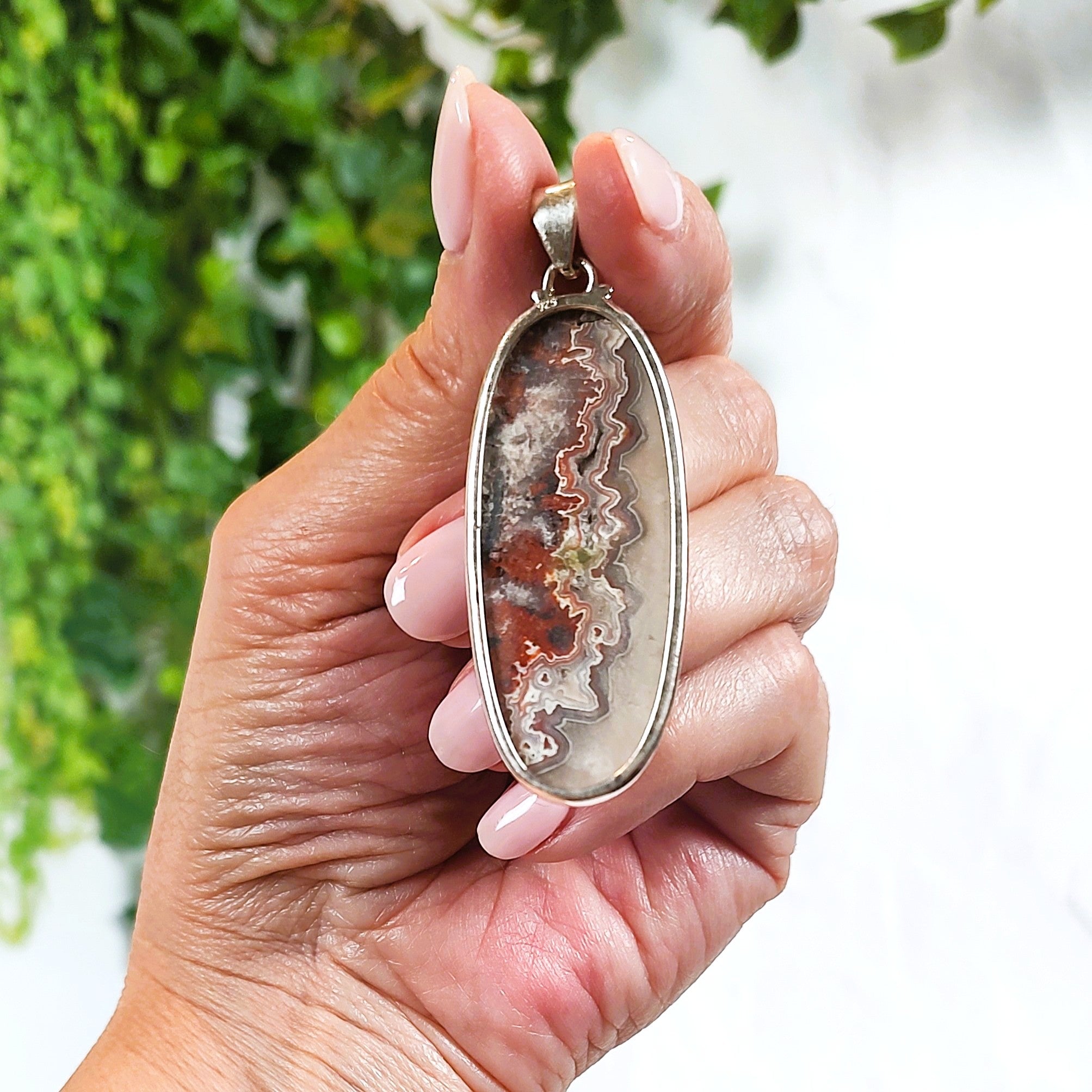 Hand holding a pendant with a natural agate stone, against a blurred green and white background.