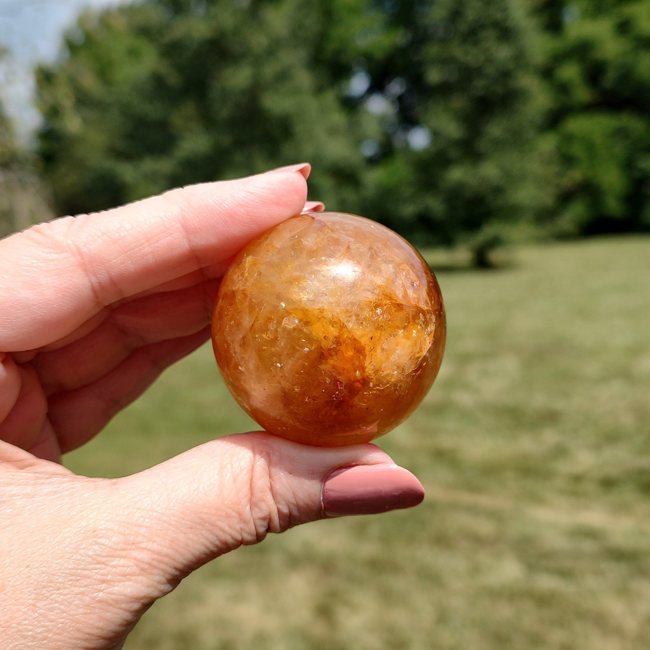Hand holding a yellow hematoid quartz sphere, outdoors in direct sunlight, with blurred background.