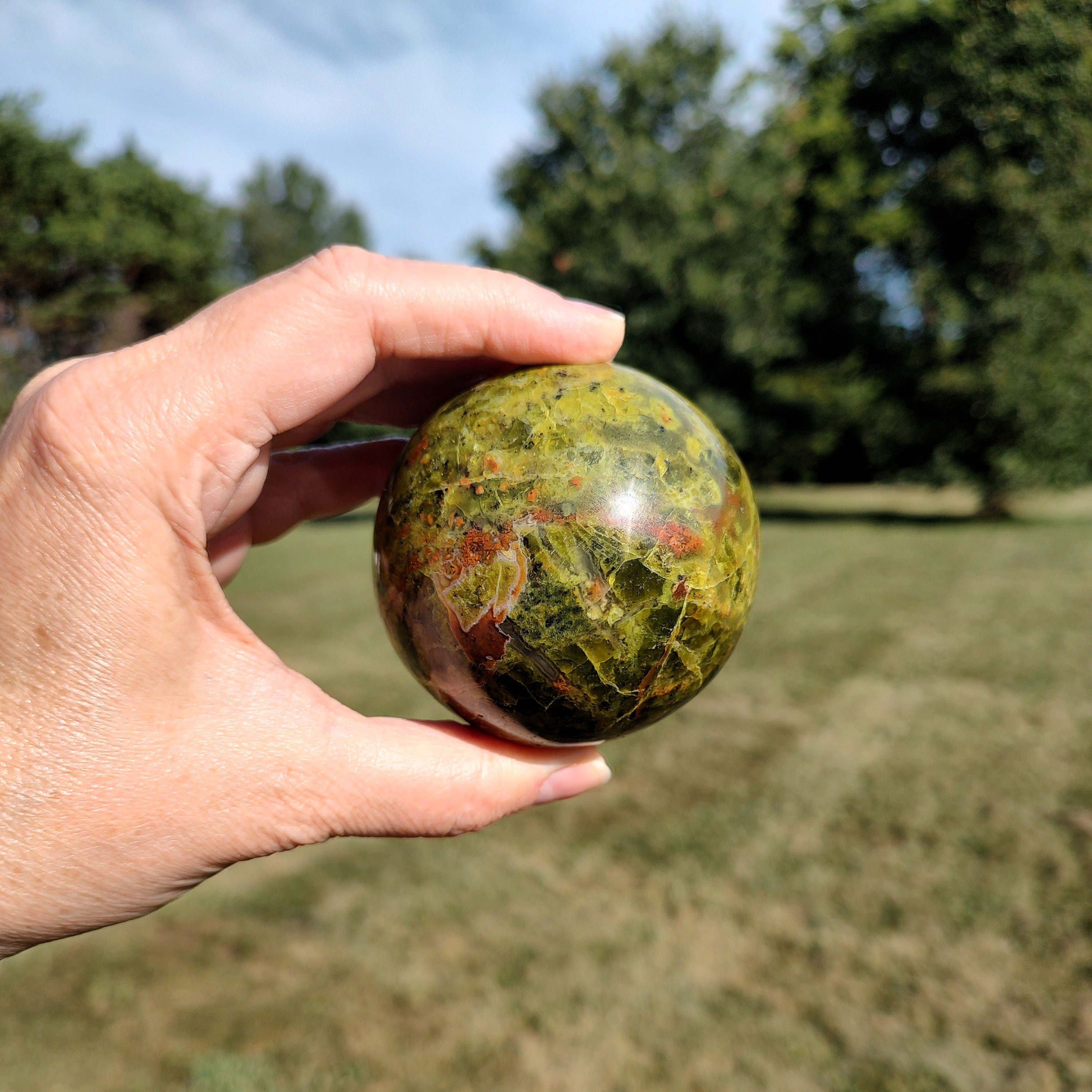 Hand holding a green opal sphere with reddish-brown marbling, against a natural outdoor background.