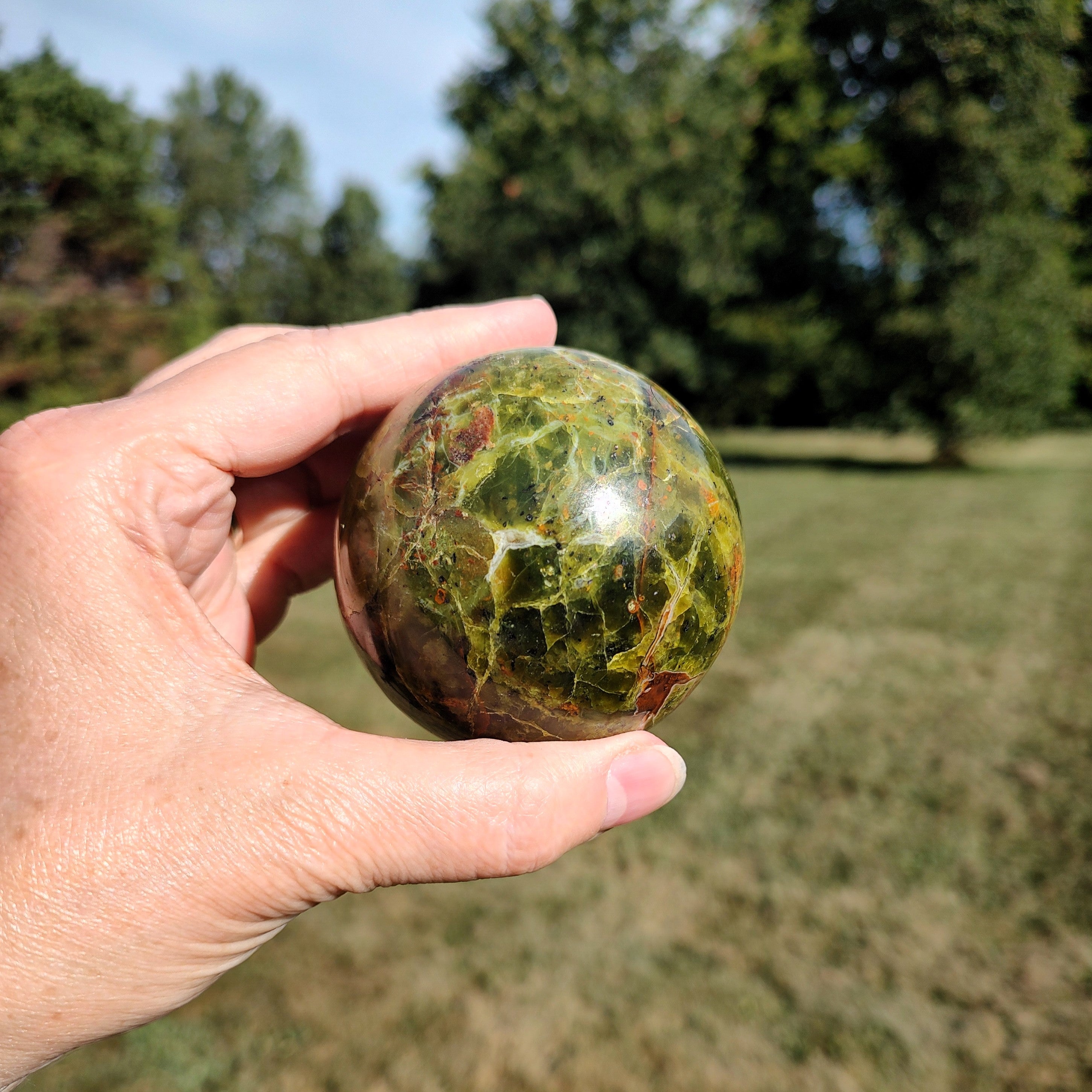 Hand holding a green and reddish-brown marbled opal sphere with a blurred natural background.