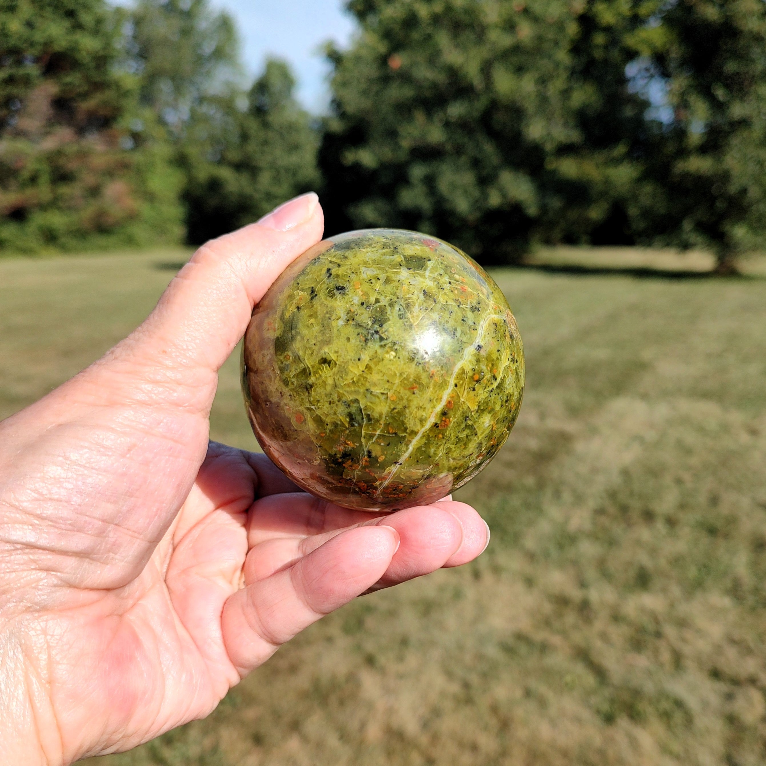 Hand holding a green opal sphere outdoors with trees in the background.