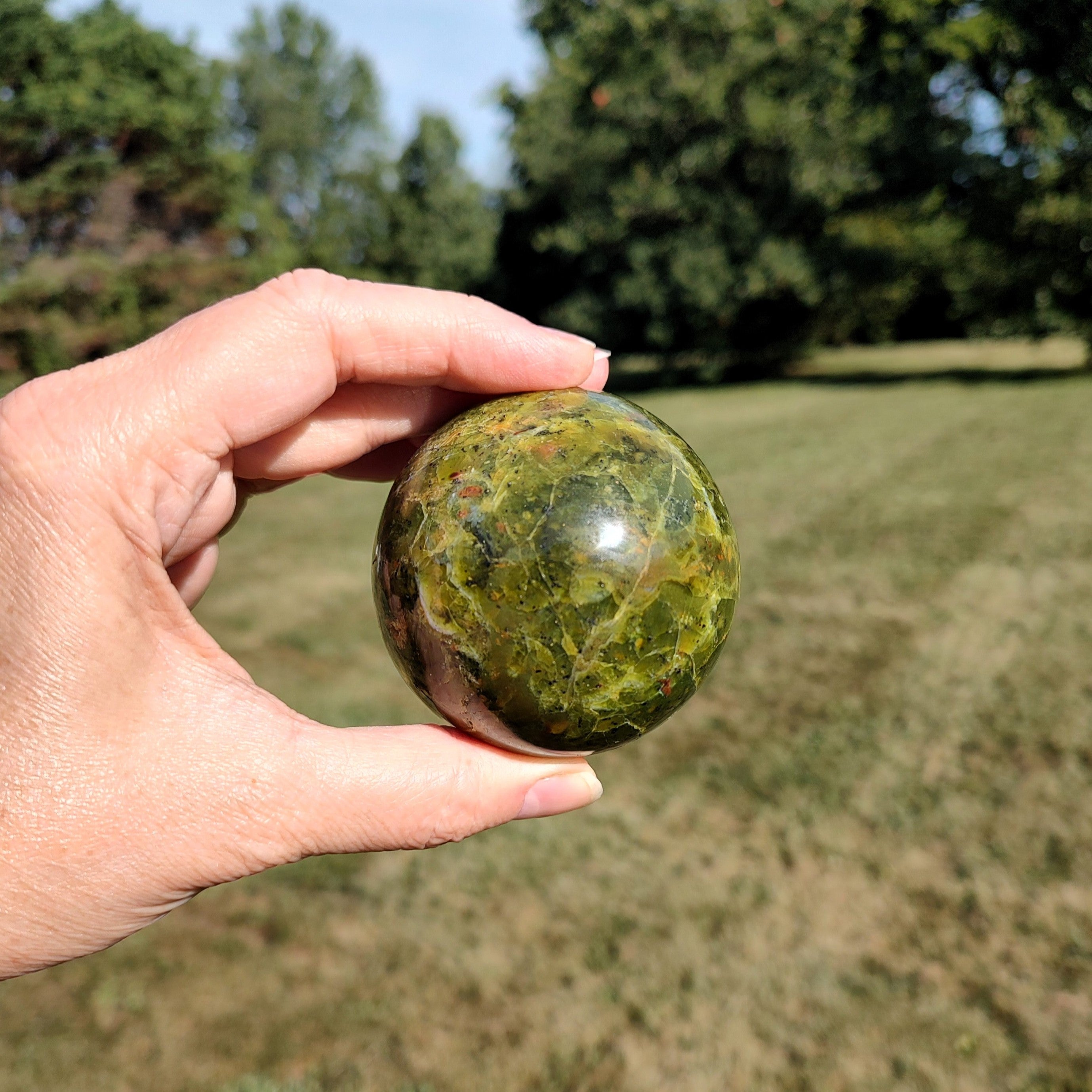 Hand holding a green opal sphere outdoors with trees in the background.