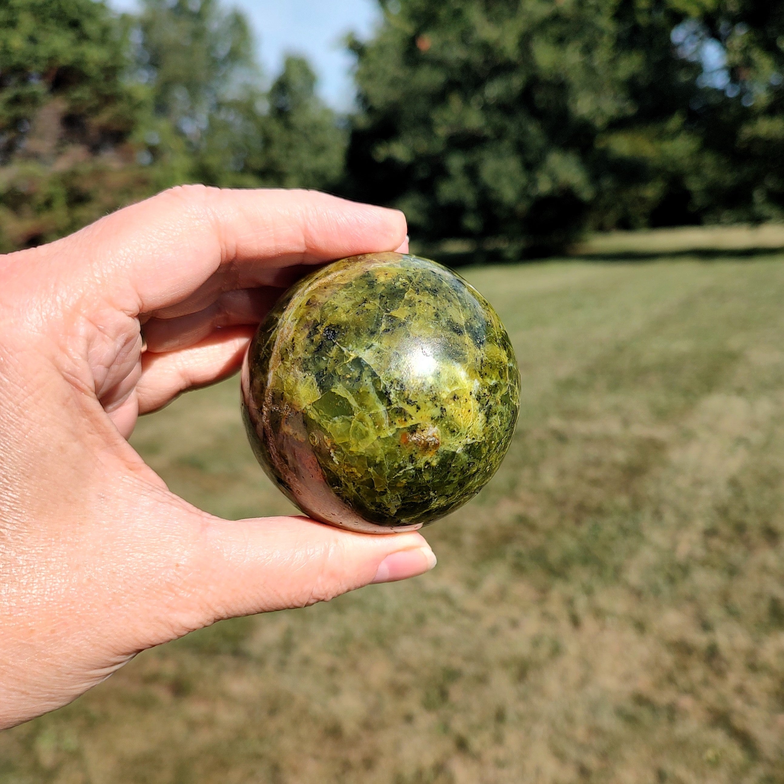 Hand holding a green opal sphere outdoors with trees in the background.