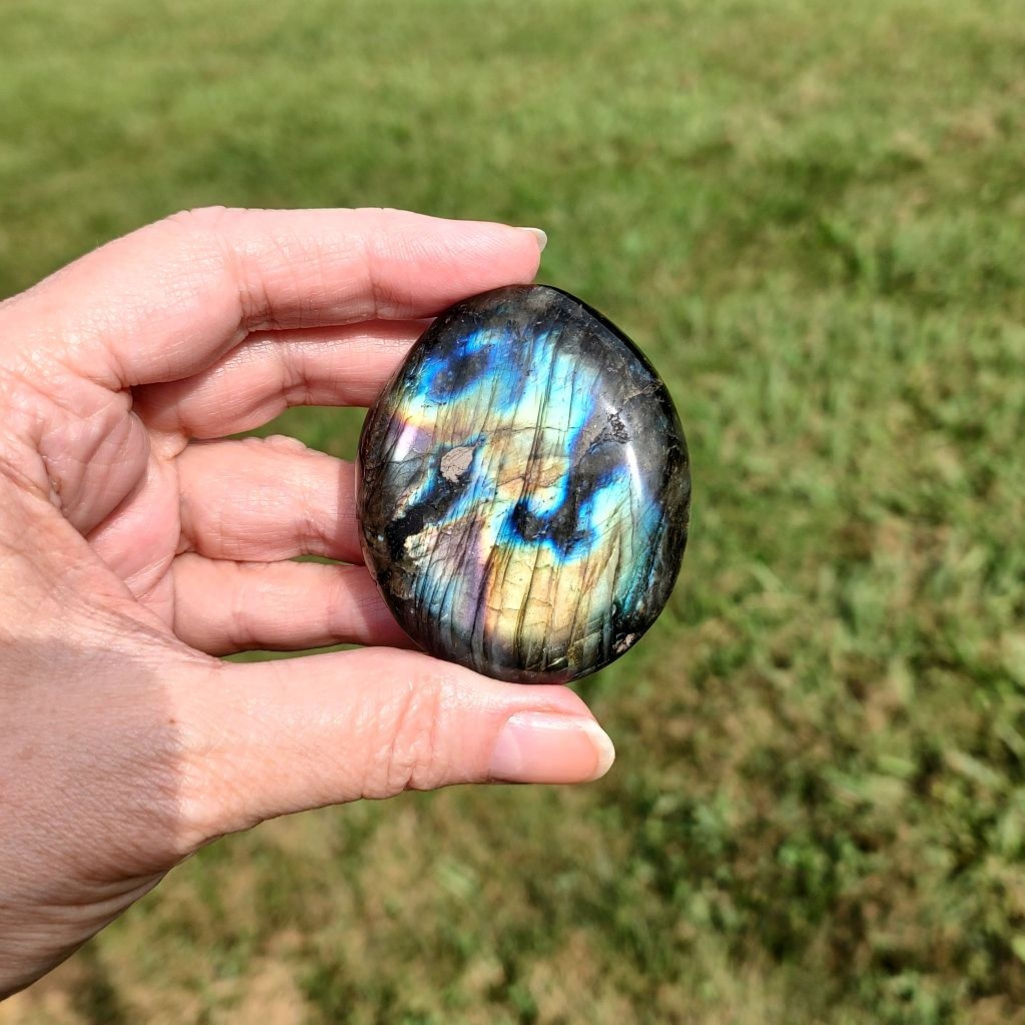 Hand holding a labradorite palm stone with a colorful pattern against a grassy background.