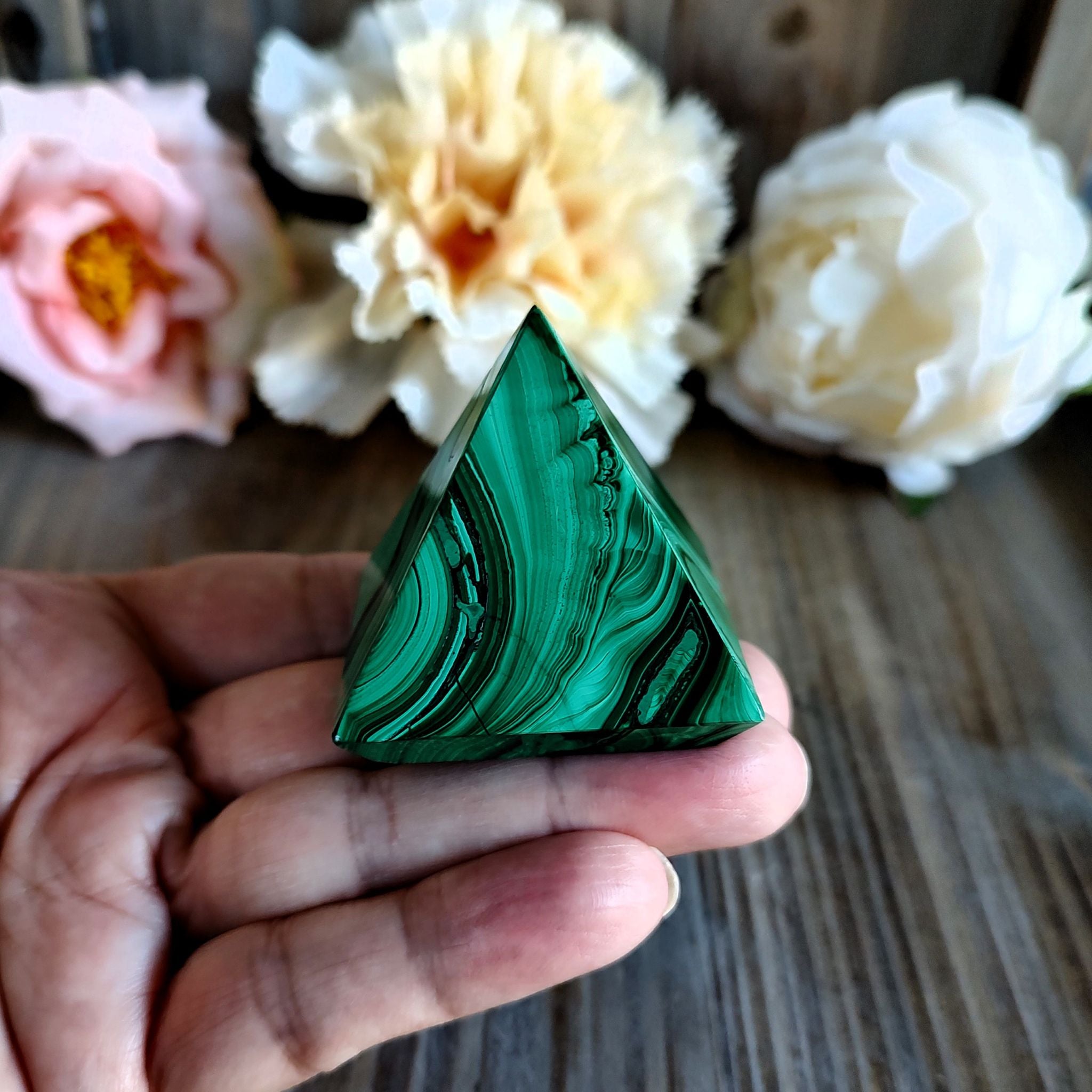 Hand holding a handcrafted malachite pyramid in front of a wooden surface with flowers in the background.