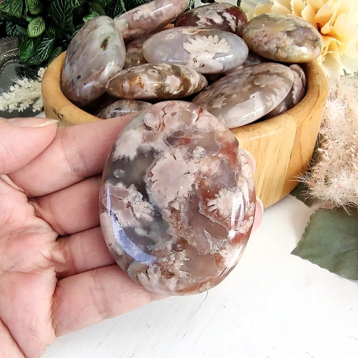 Polished flower agate palm stone held in a hand, with a bowl of additional stones in the background.