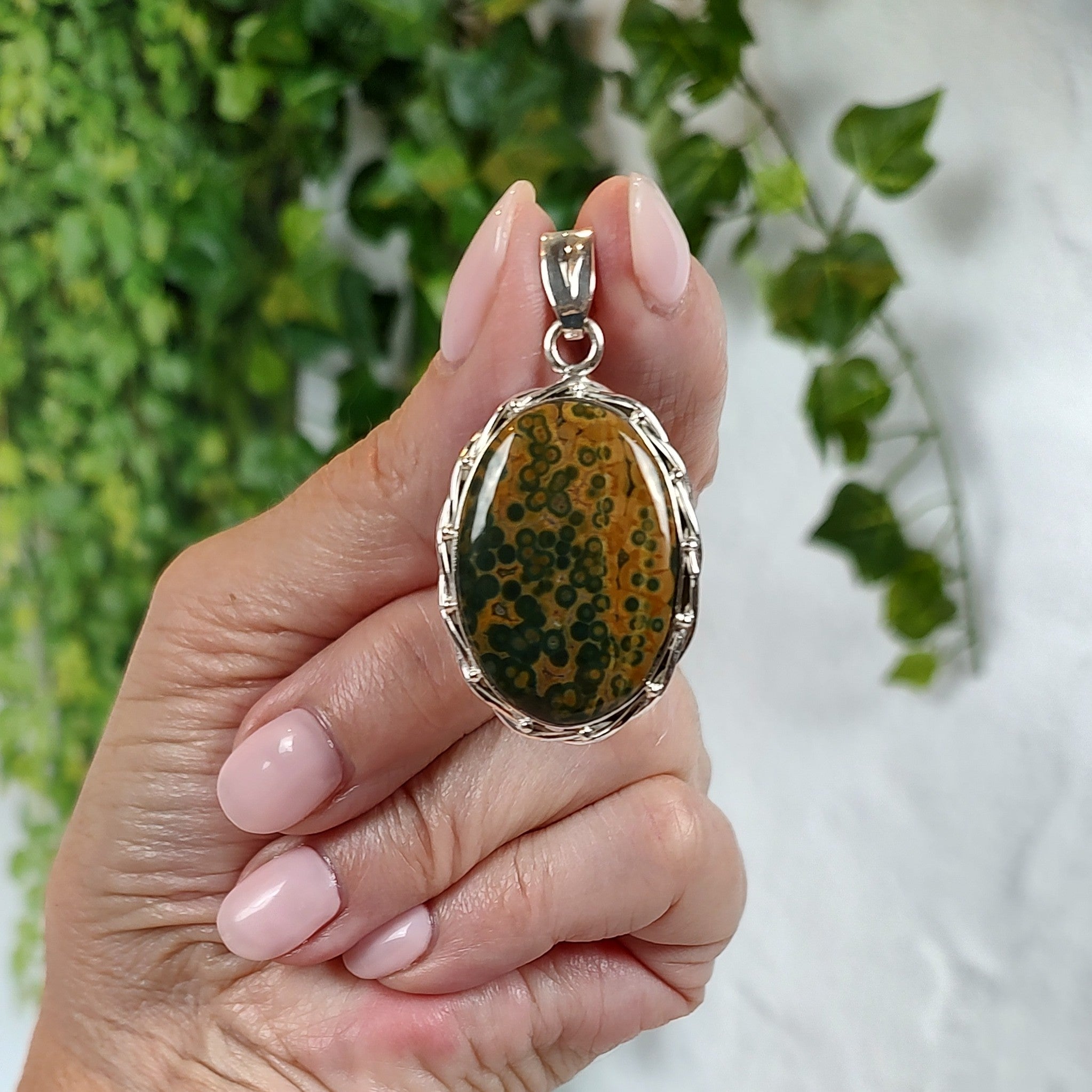 Hand holding a silver pendant with a green and golden brown stone, against a blurred natural background.