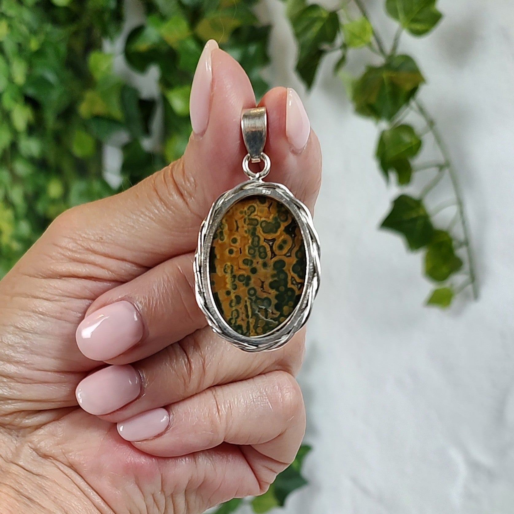 Hand holding a silver and ocean jasper pendant against a blurred green and white background.