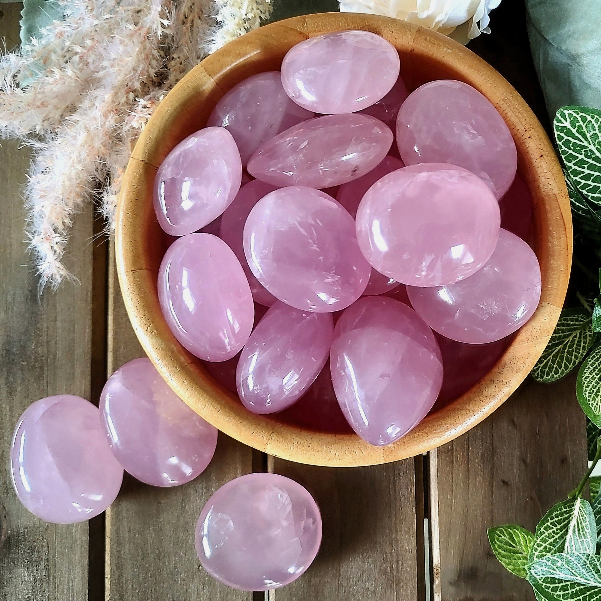 Polished rose quartz pebbles in a wooden bowl, surrounded by leaves and flowers on a wooden surface.