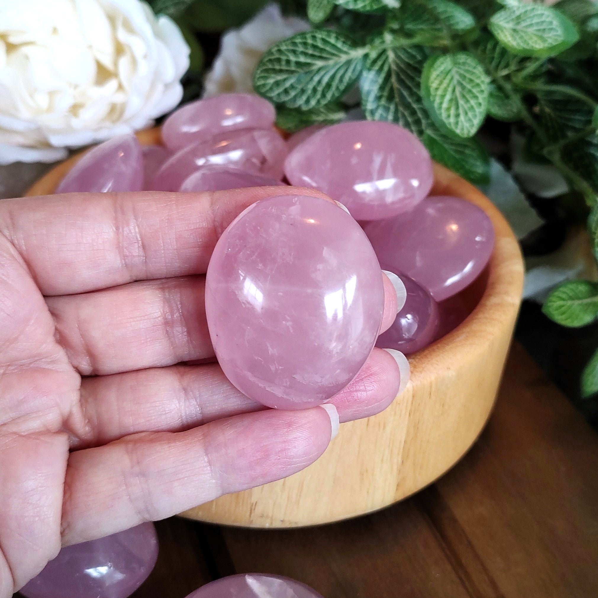A polished rose quartz pebble shown in a hand, with green leaves, white flowers, and a wooden bowl containing rose quartz pebbles.