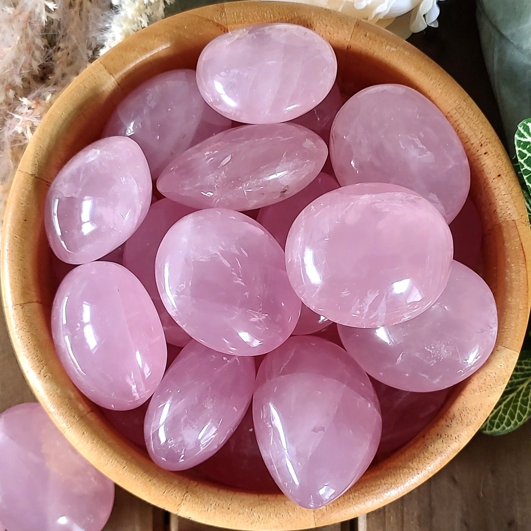 Polished rose quartz pebbles in a wooden bowl, with leaves and flowers in the photo edges.