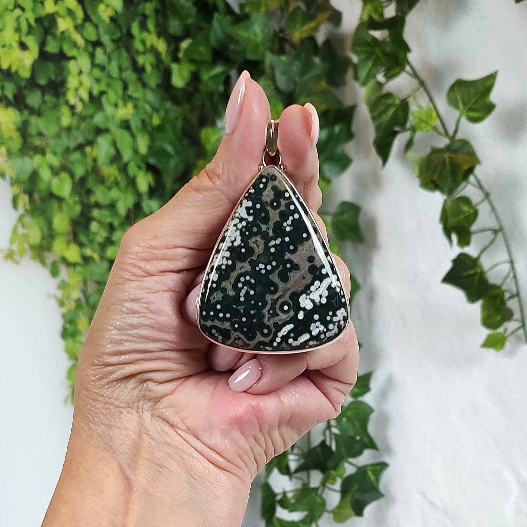 Hand holding a large ocean jasper pendant with greenery in the background.