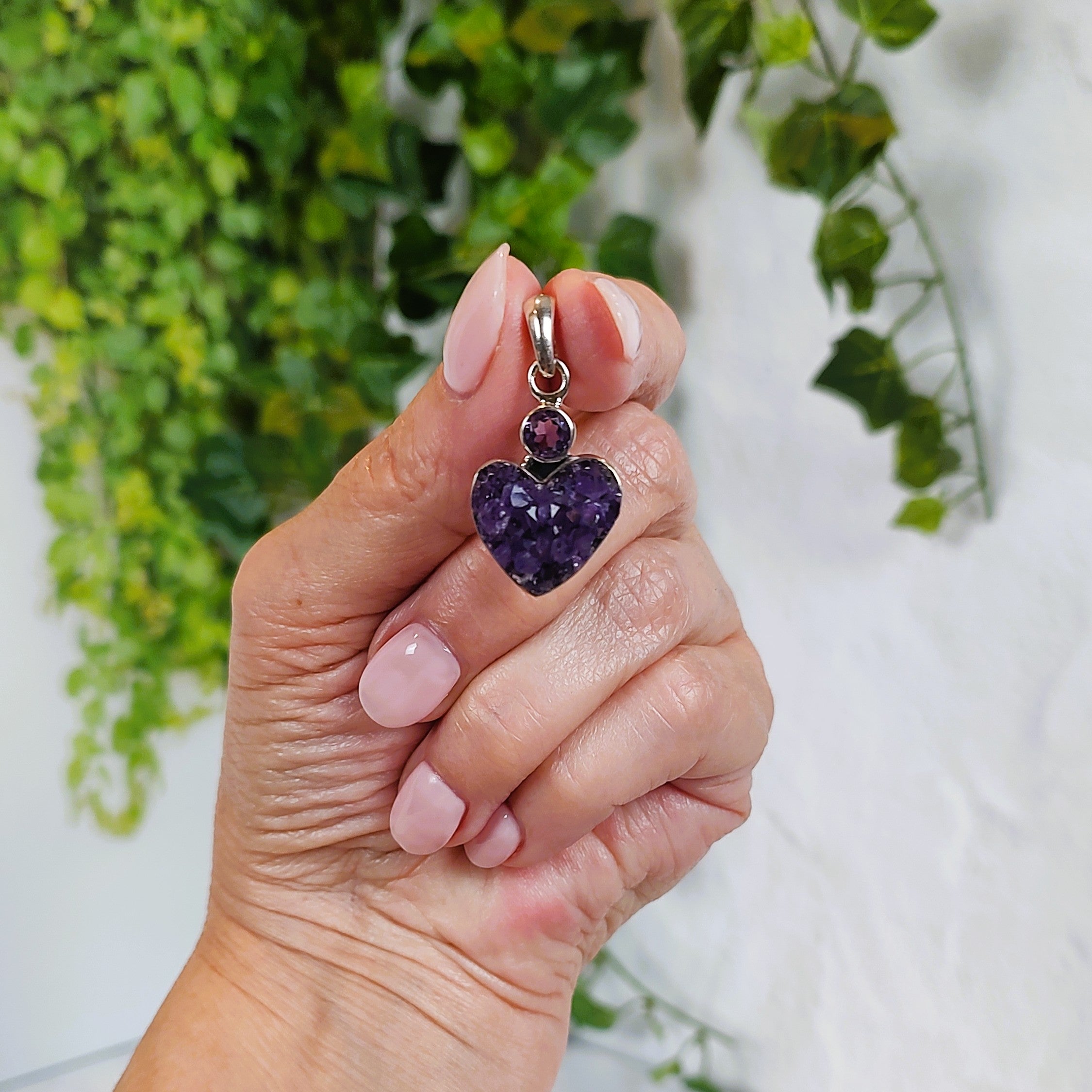 Hand holding a heart-shaped amethyst cluster pendant with greenery in the background.