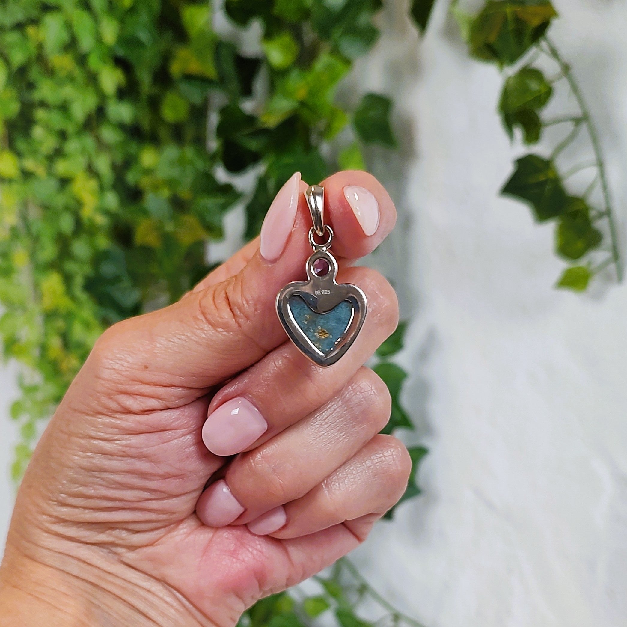 Hand holding a heart-shaped pendant, showing the back, against a blurred green and white background.