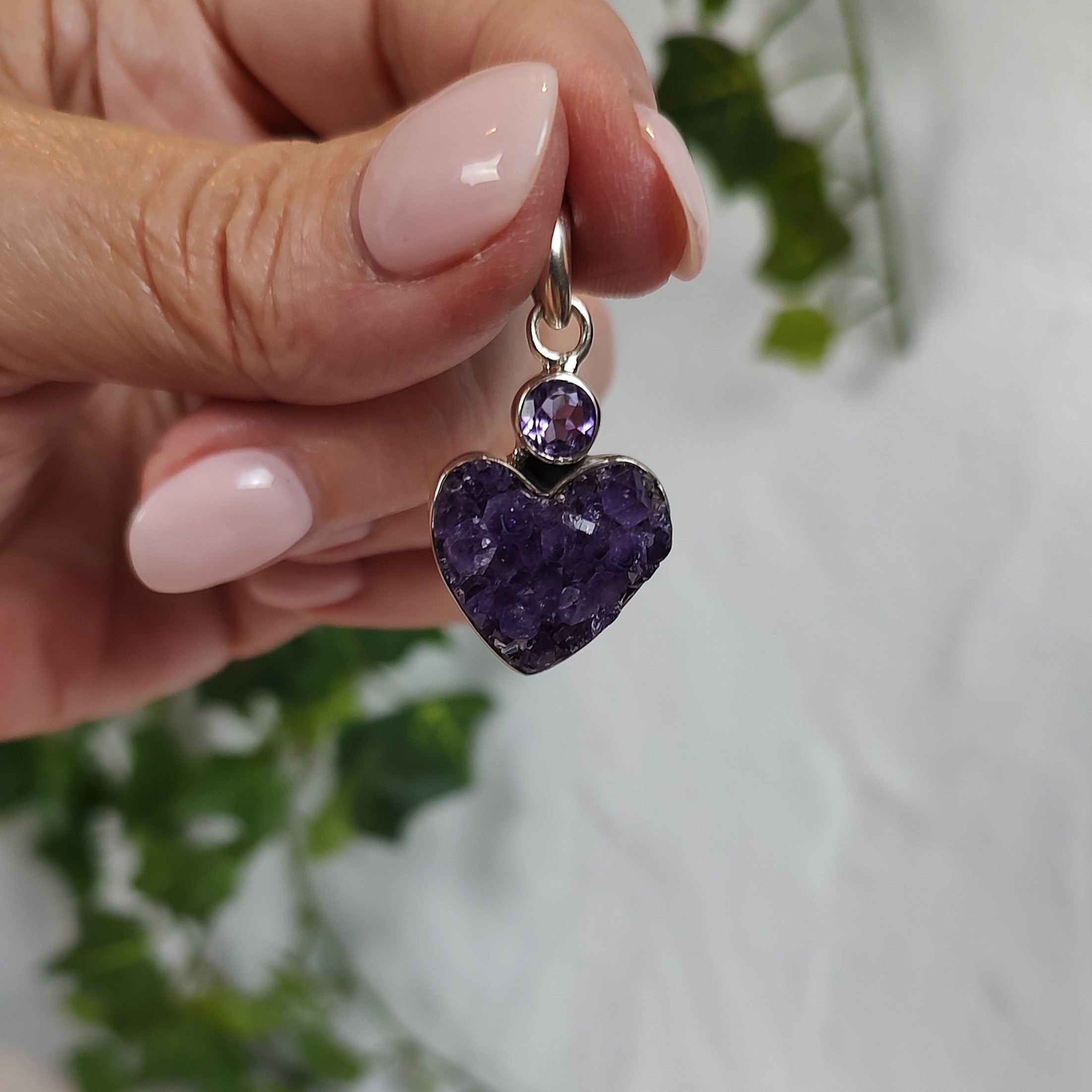 Hand holding a heart-shaped amethyst cluster pendant with a blurred green leafy background.