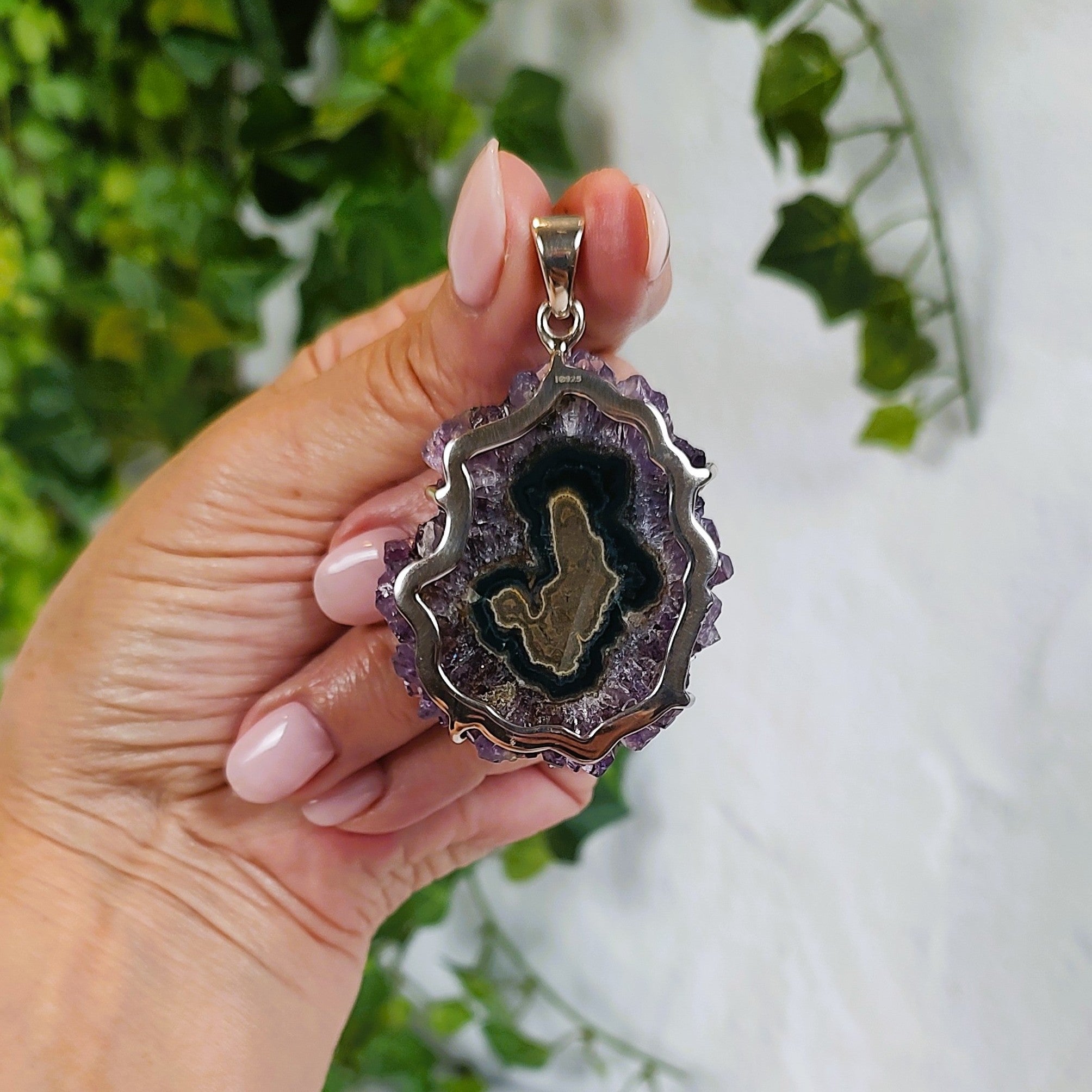 Hand holding an amethyst and agate pendant, showing the back, with a blurred green and white background,