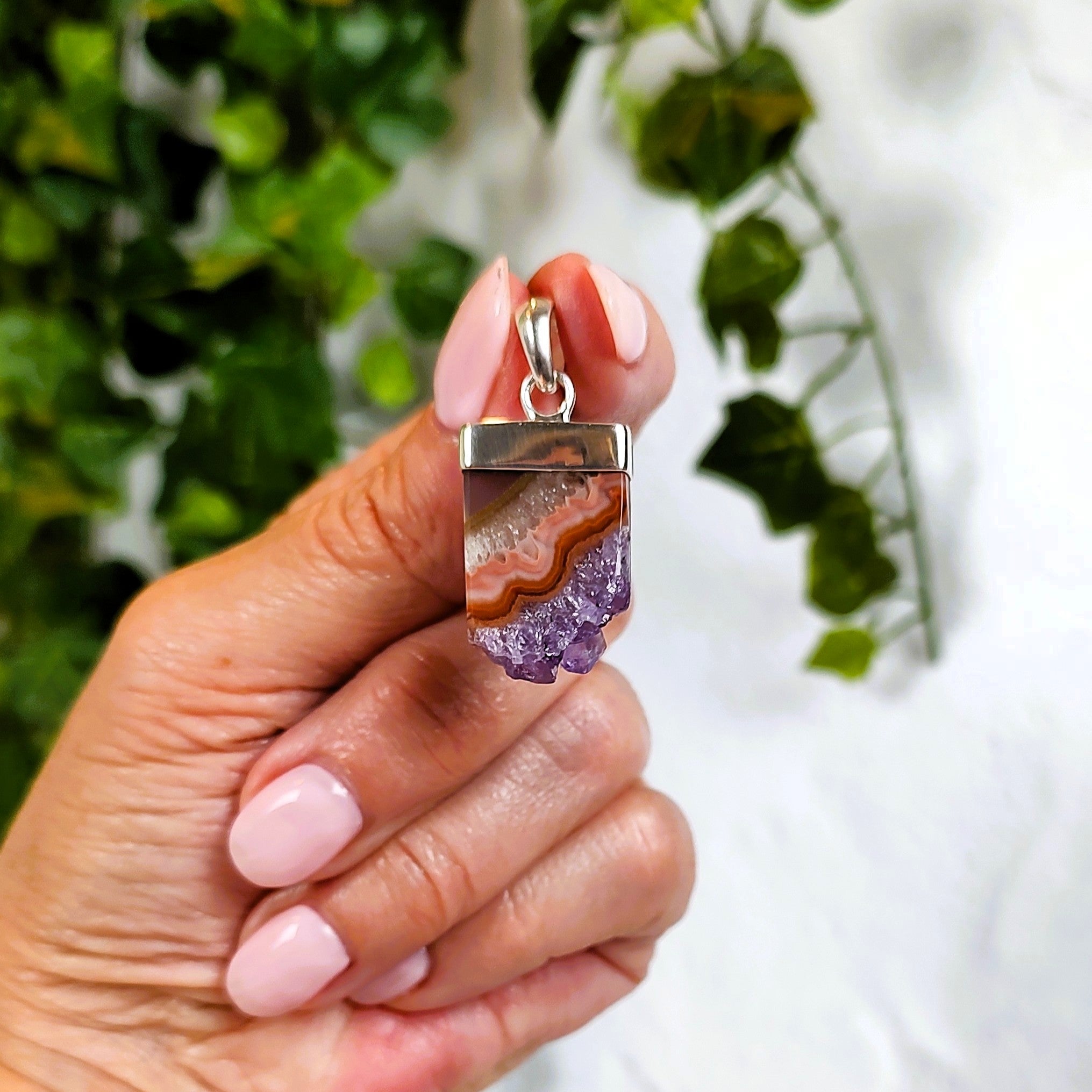 Hand holding a amethyst and agate pendant with a blurred green and white background.