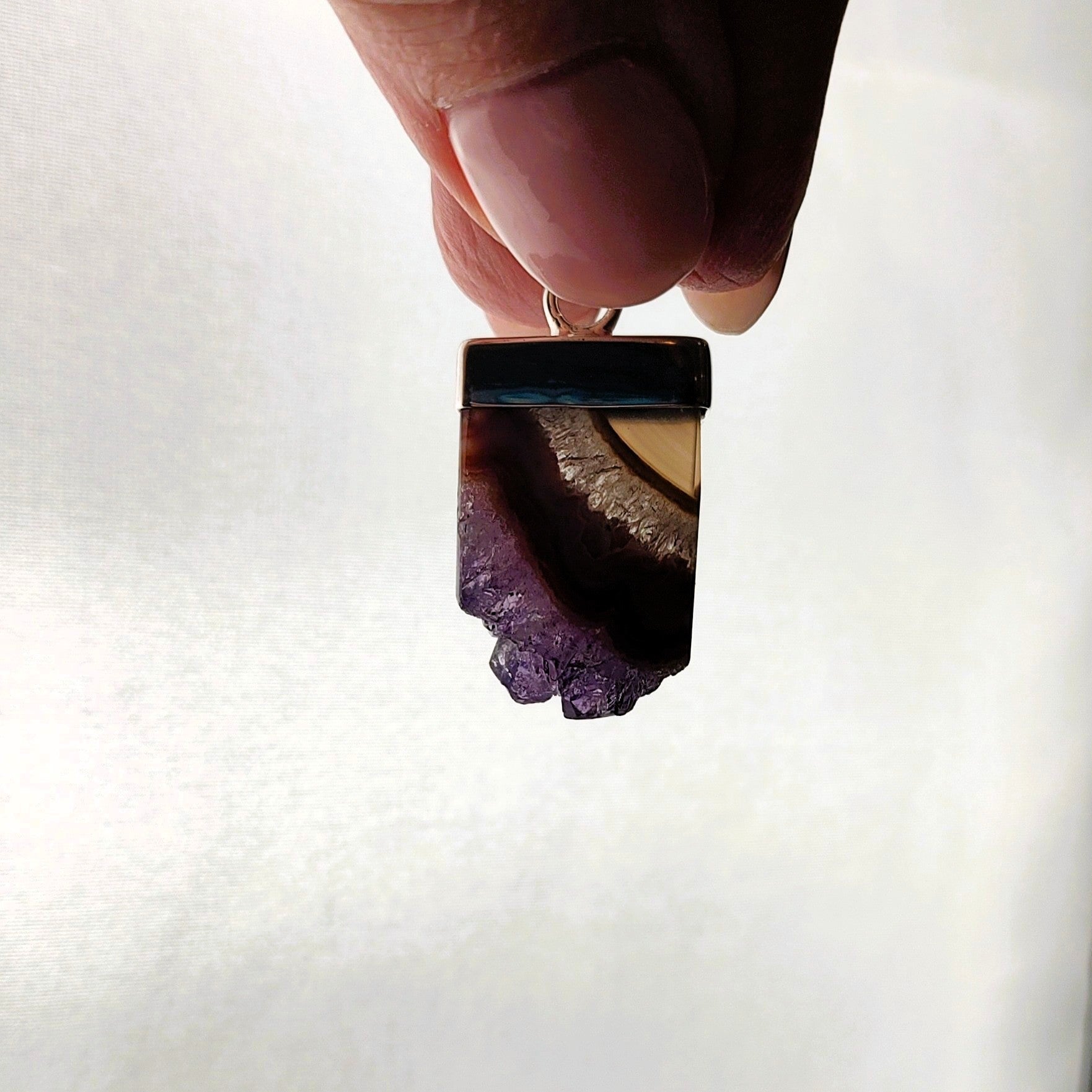 Hand holding a backlit amethyst and agate pendant against a white background.