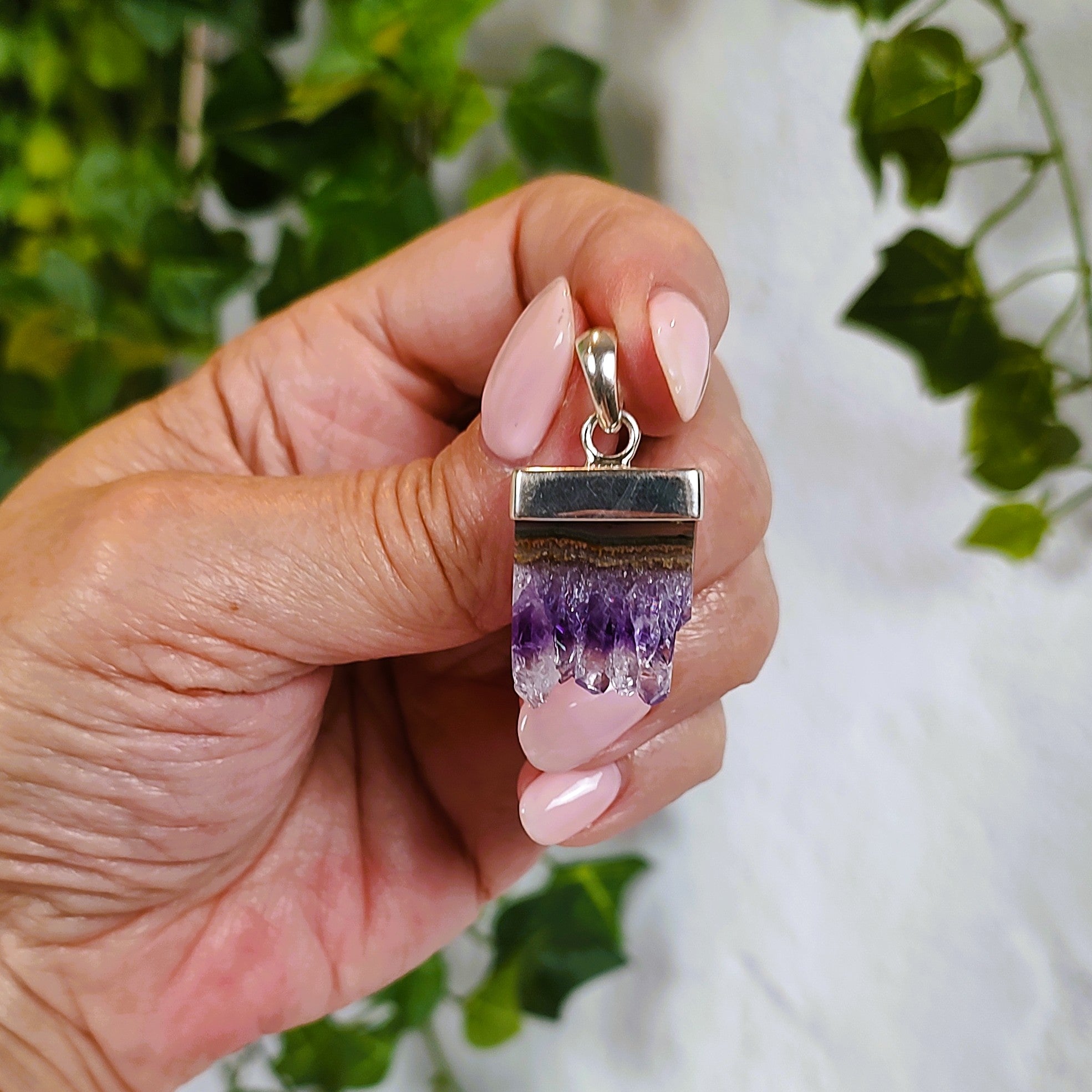 Hand holding a silver pendant with an amethyst and agate stalactite slice, against a blurred green and white background.  