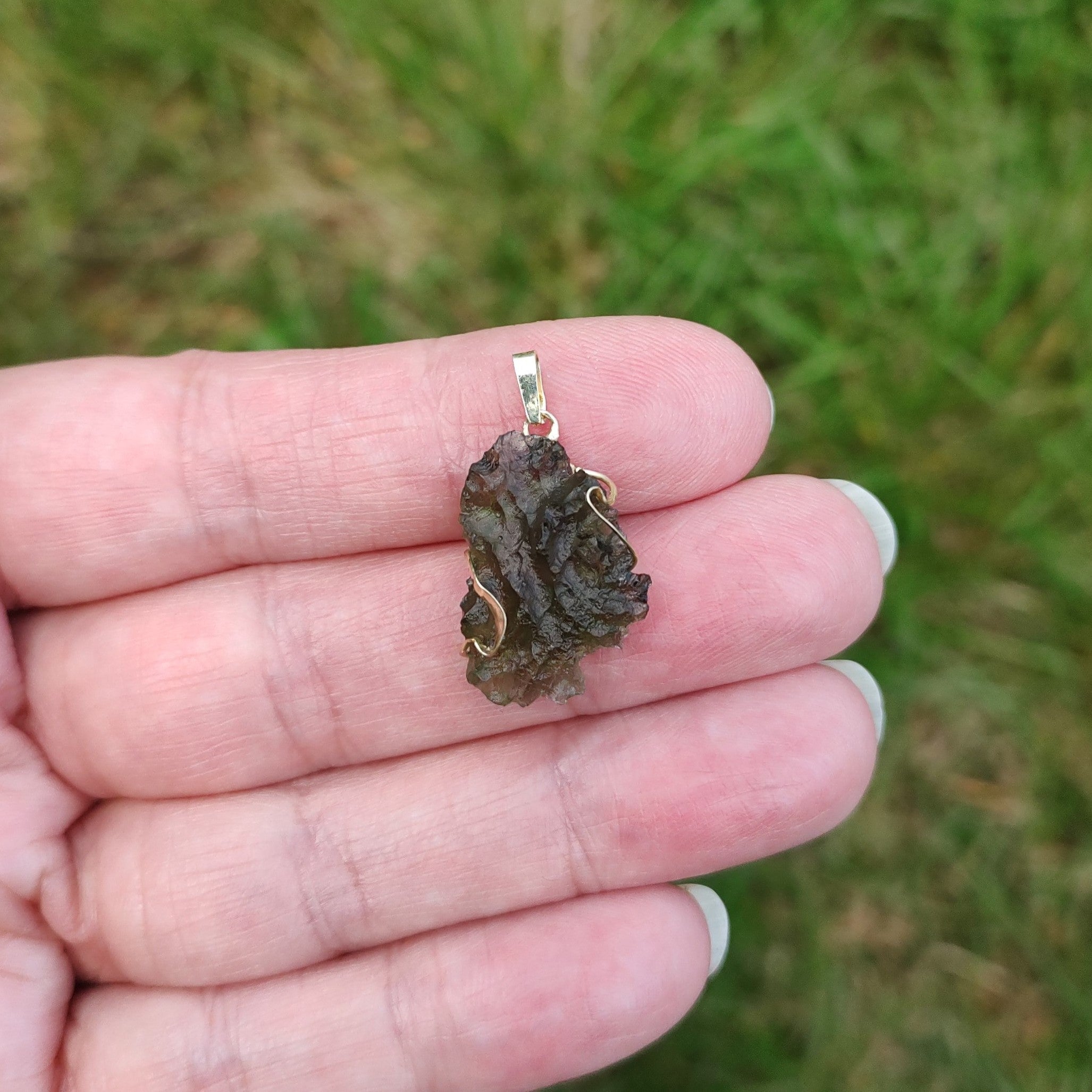 Hand holding a raw moldavite pendant set in gold, against a grassy background.
