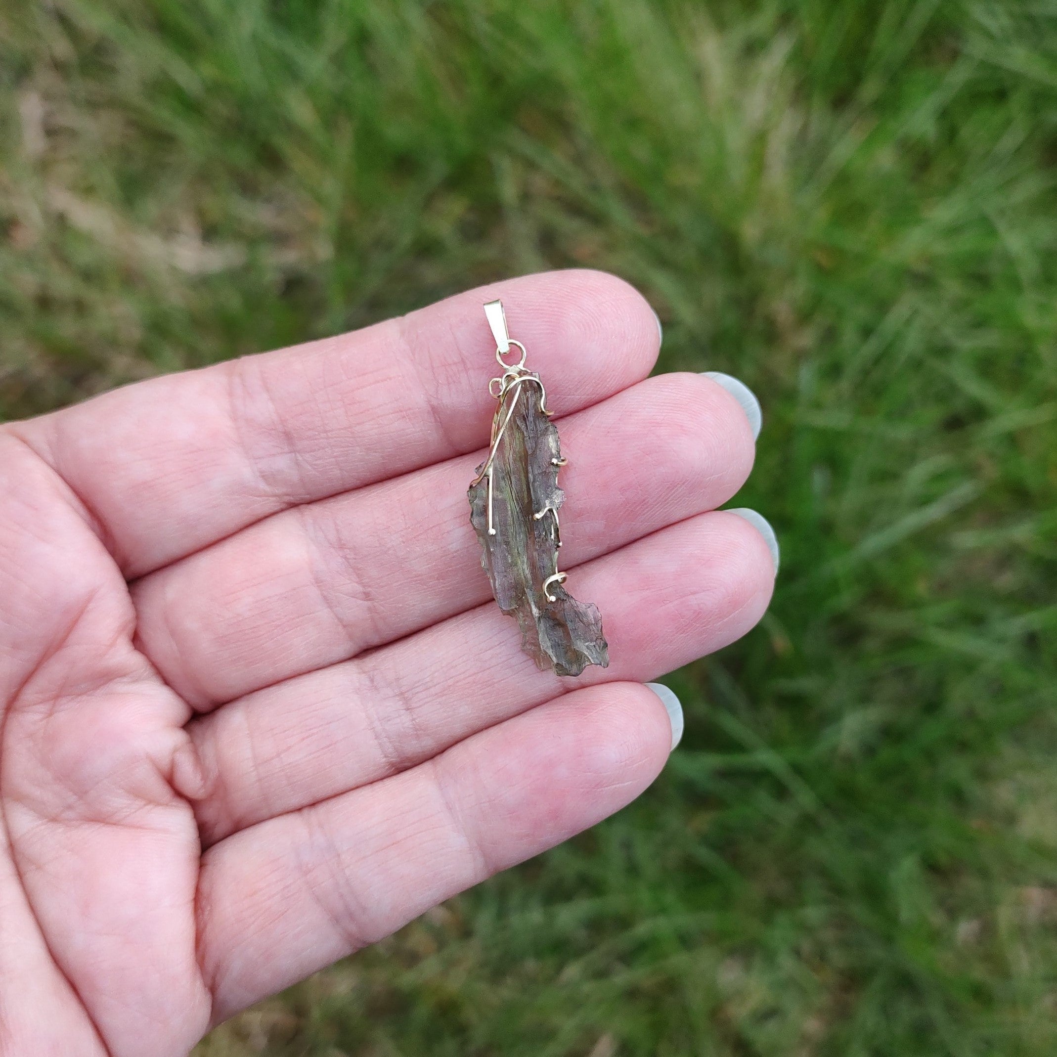 Hand holding a raw moldavite pendant set in gold, against a grassy background.
