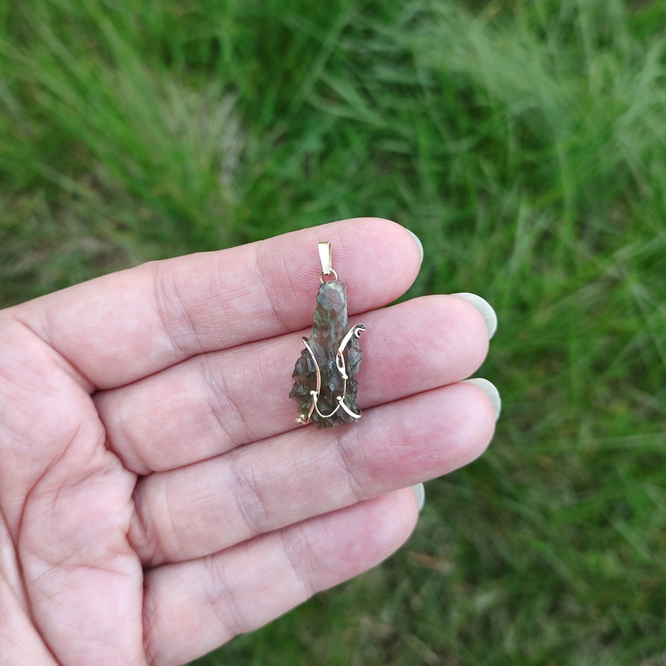Hand holding a raw moldavite pendant set in gold, against a grassy background.
