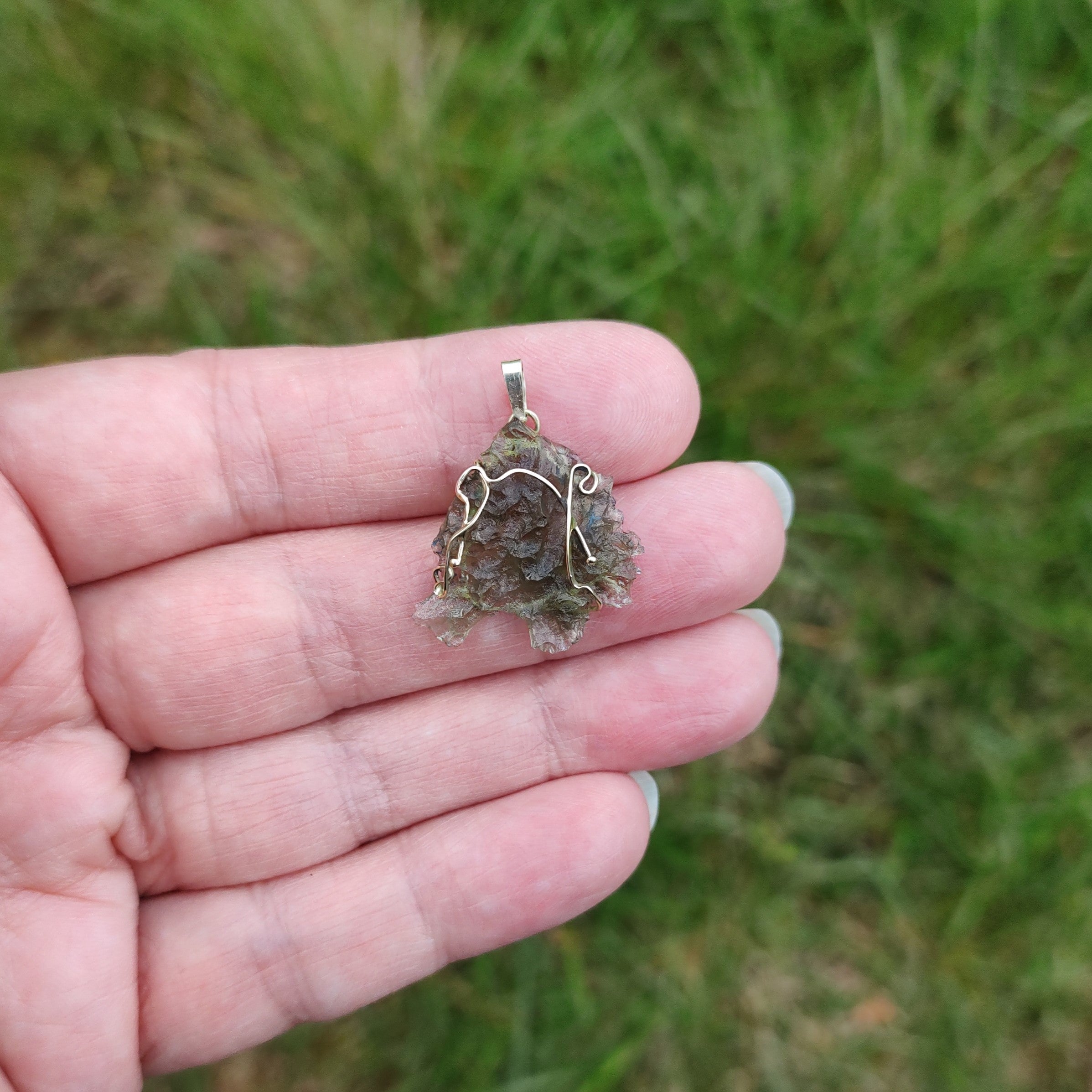 Hand holding a raw moldavite pendant set in gold, against a grassy background.