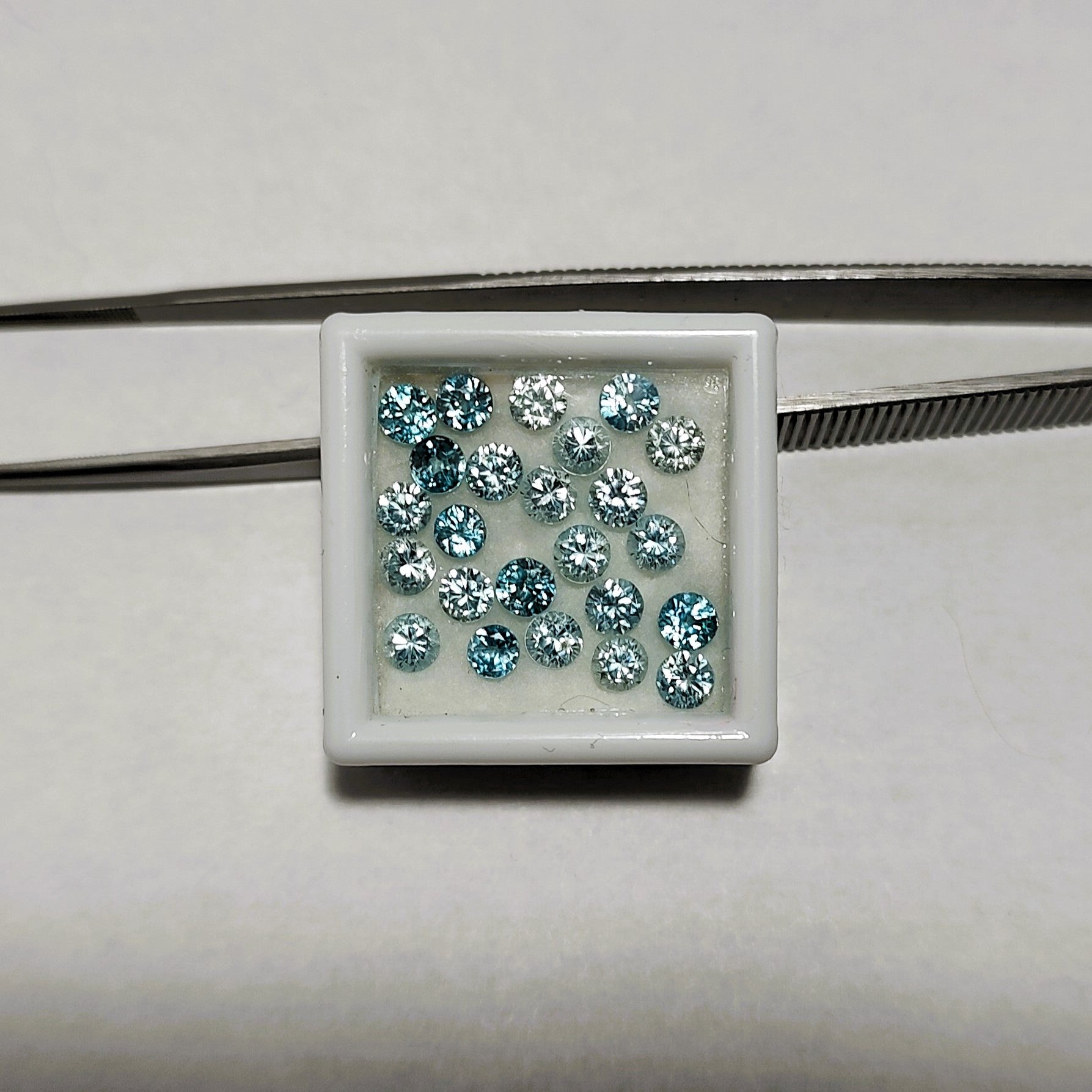 A parcel of faceted round blue zircons is shown in a gem jar, with metal tweezers in the background. 