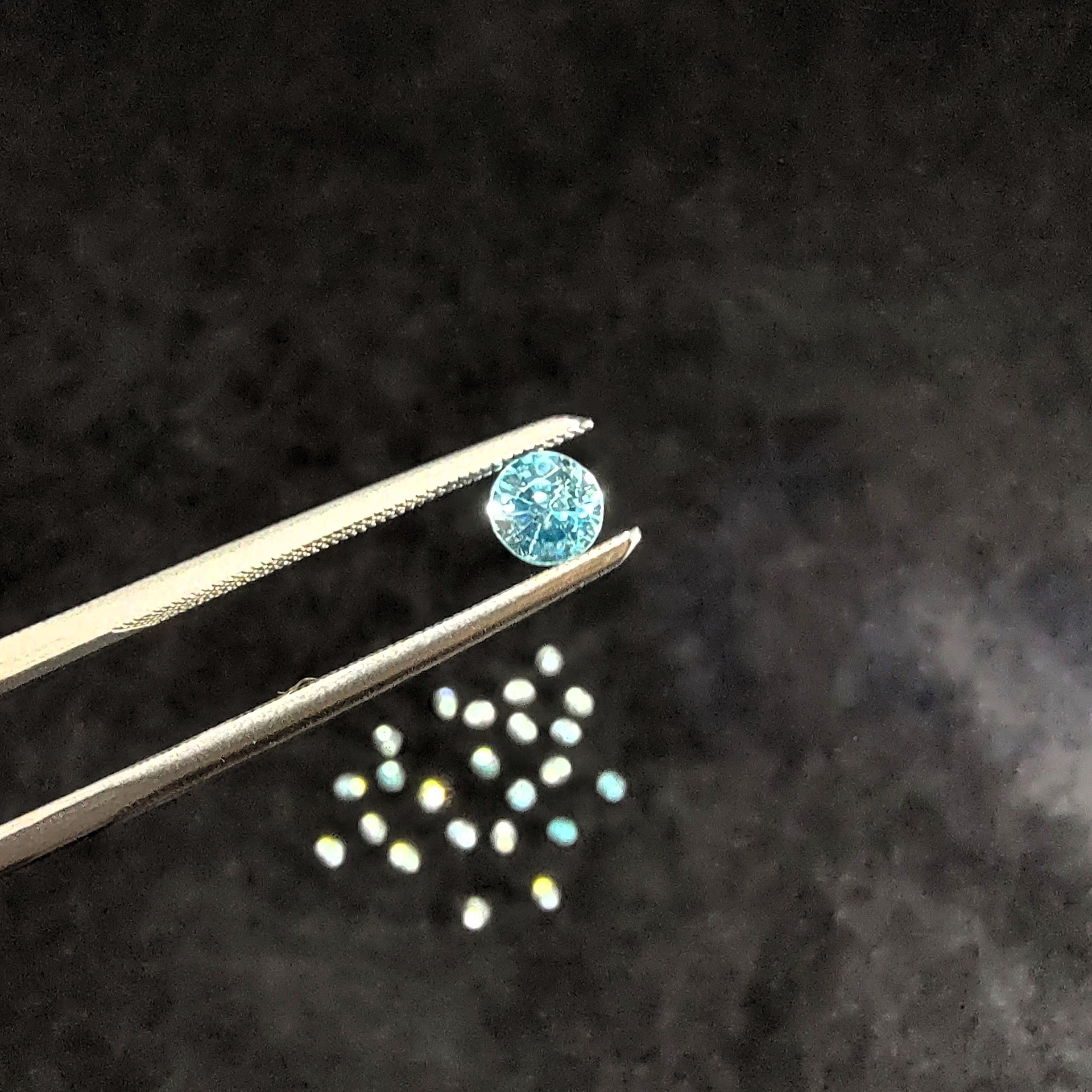 Round faceted blue zircon, held by tweezers against a gray background.