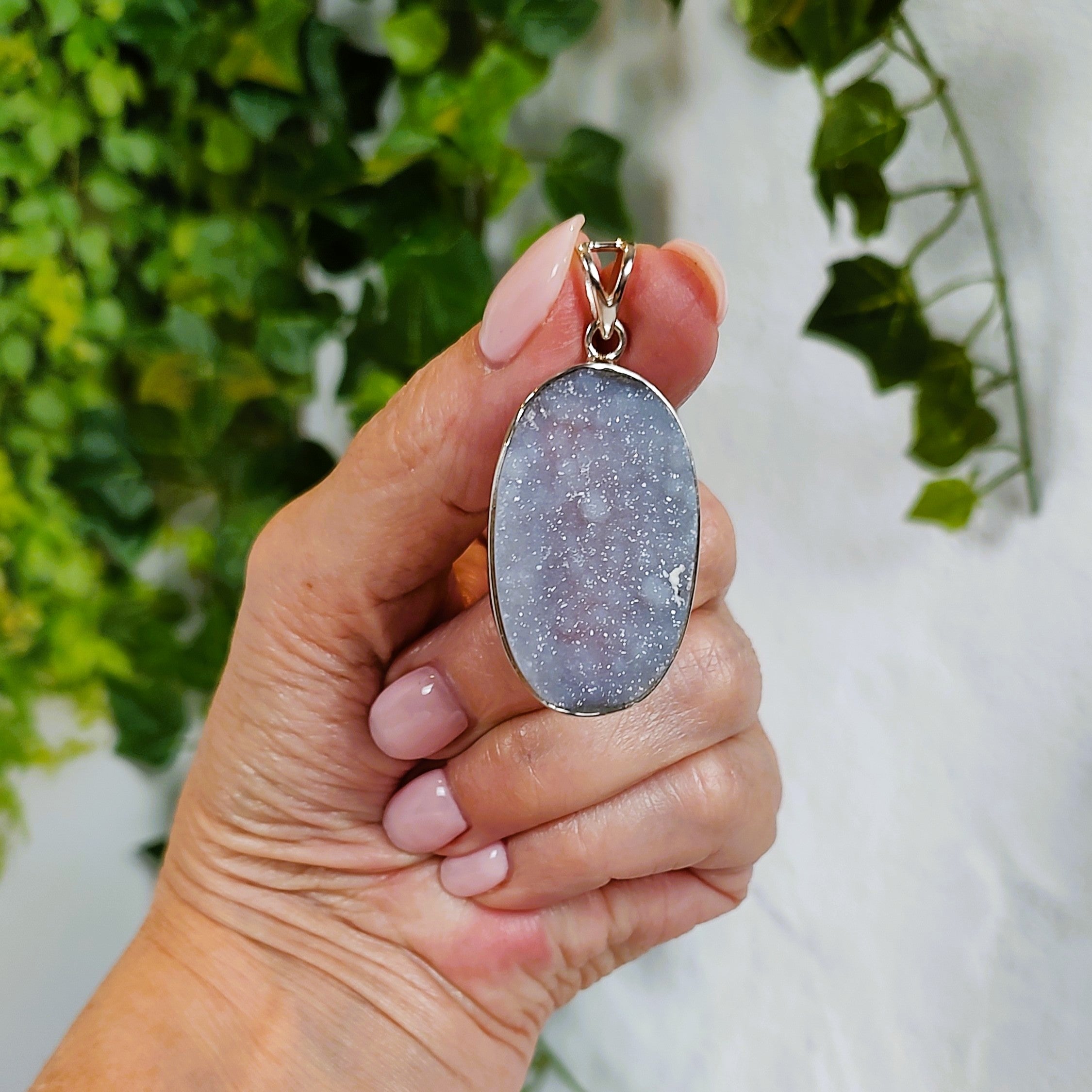 Hand holding a pendant with a grayish blue chalcedony stone against a blurred green and white background.