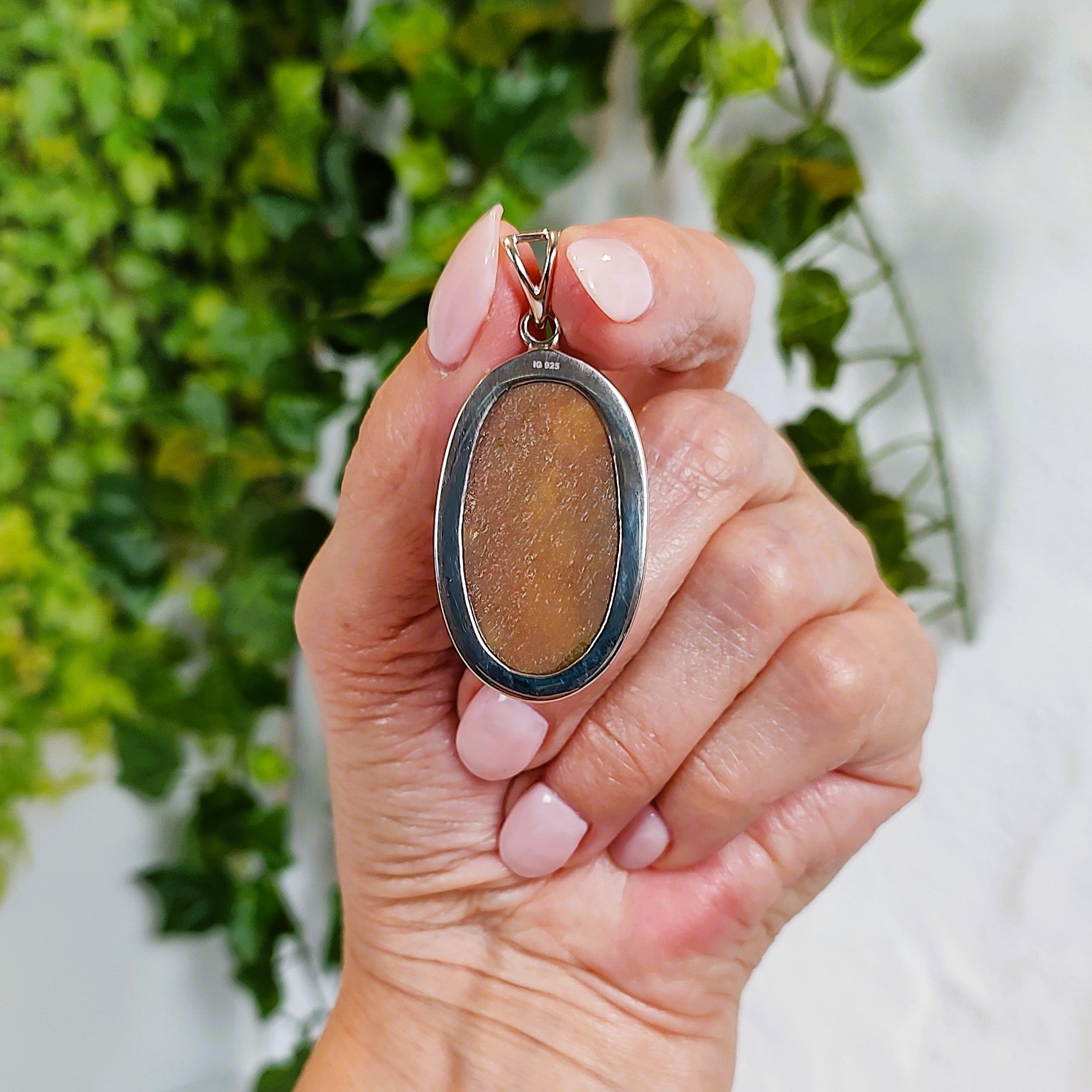 Hand holding a pendant, showing the back of the piece, with a brown oval stone against a green leafy background.