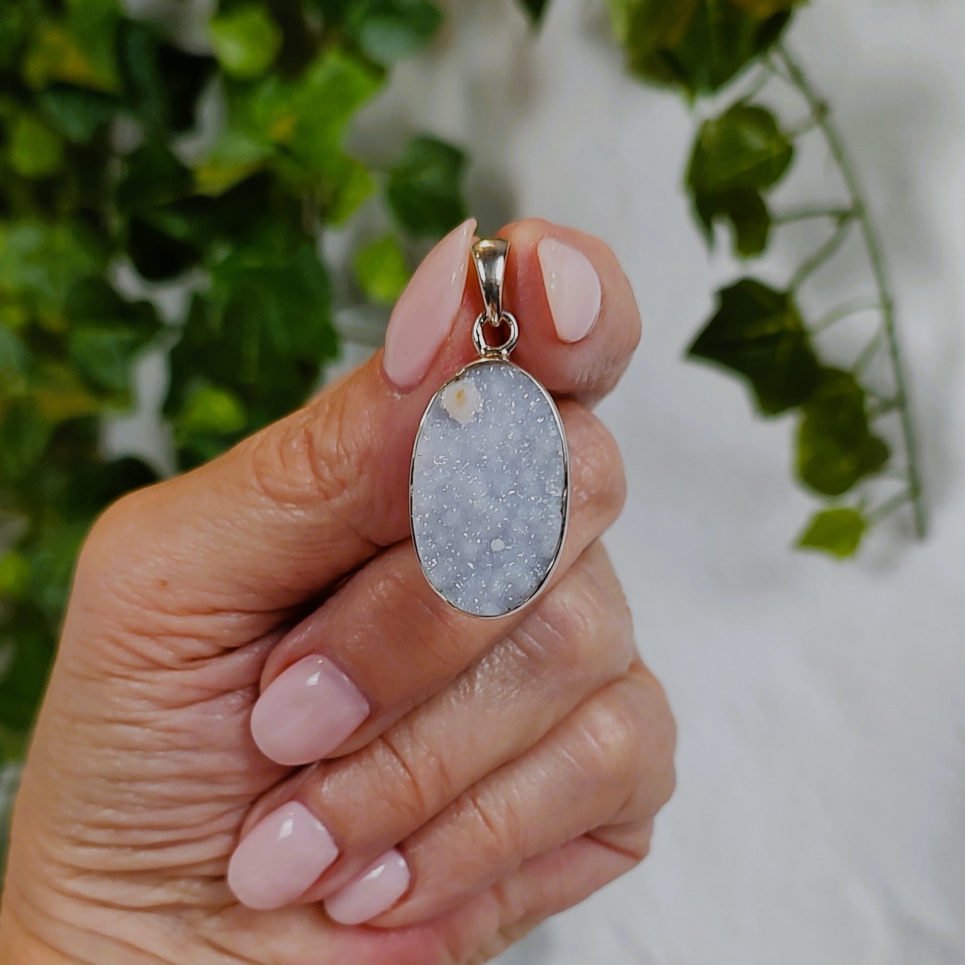Hand holding a pendant with a grayish blue chalcedony stone against a blurred green and white background.