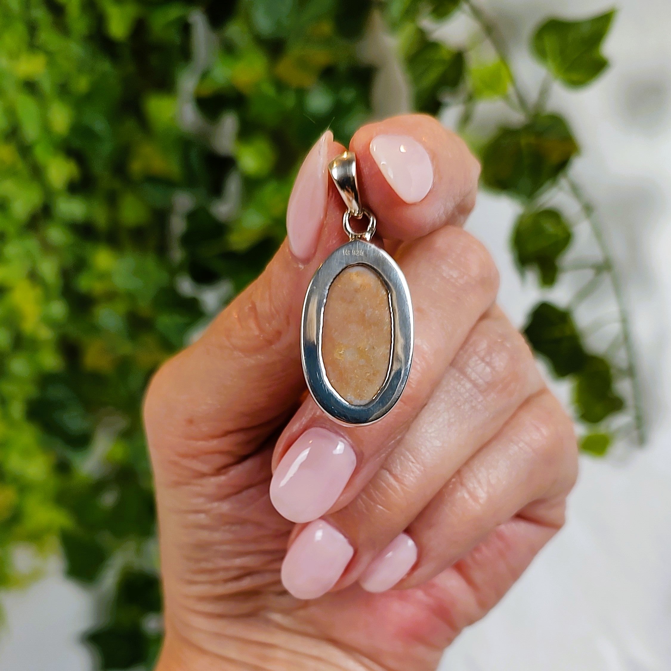 Hand holding a pendant, showing the back of the piece, with a brown oval stone against a green leafy background.