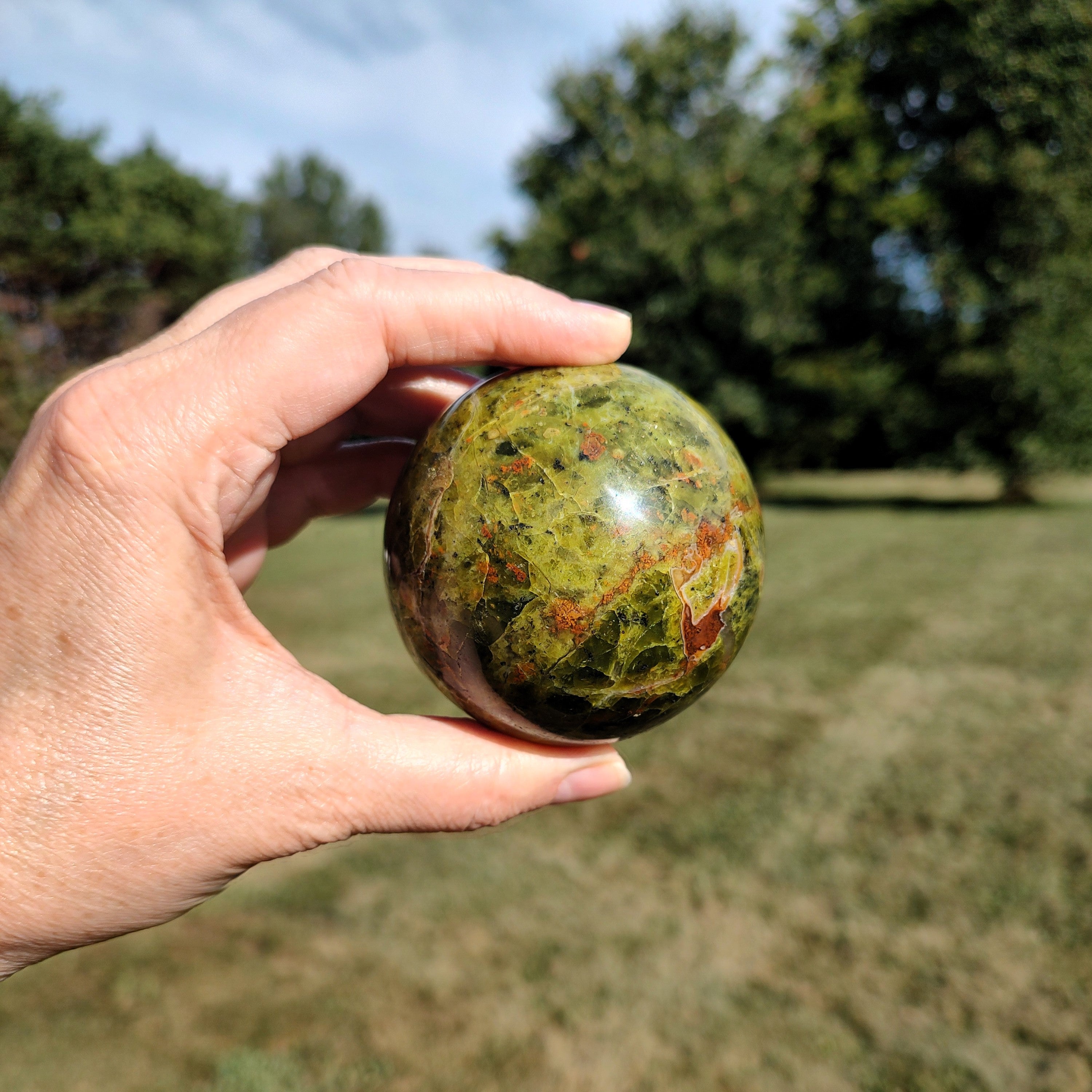 Hand holding a green opal sphere with reddish-brown marbling, against a natural outdoor background.